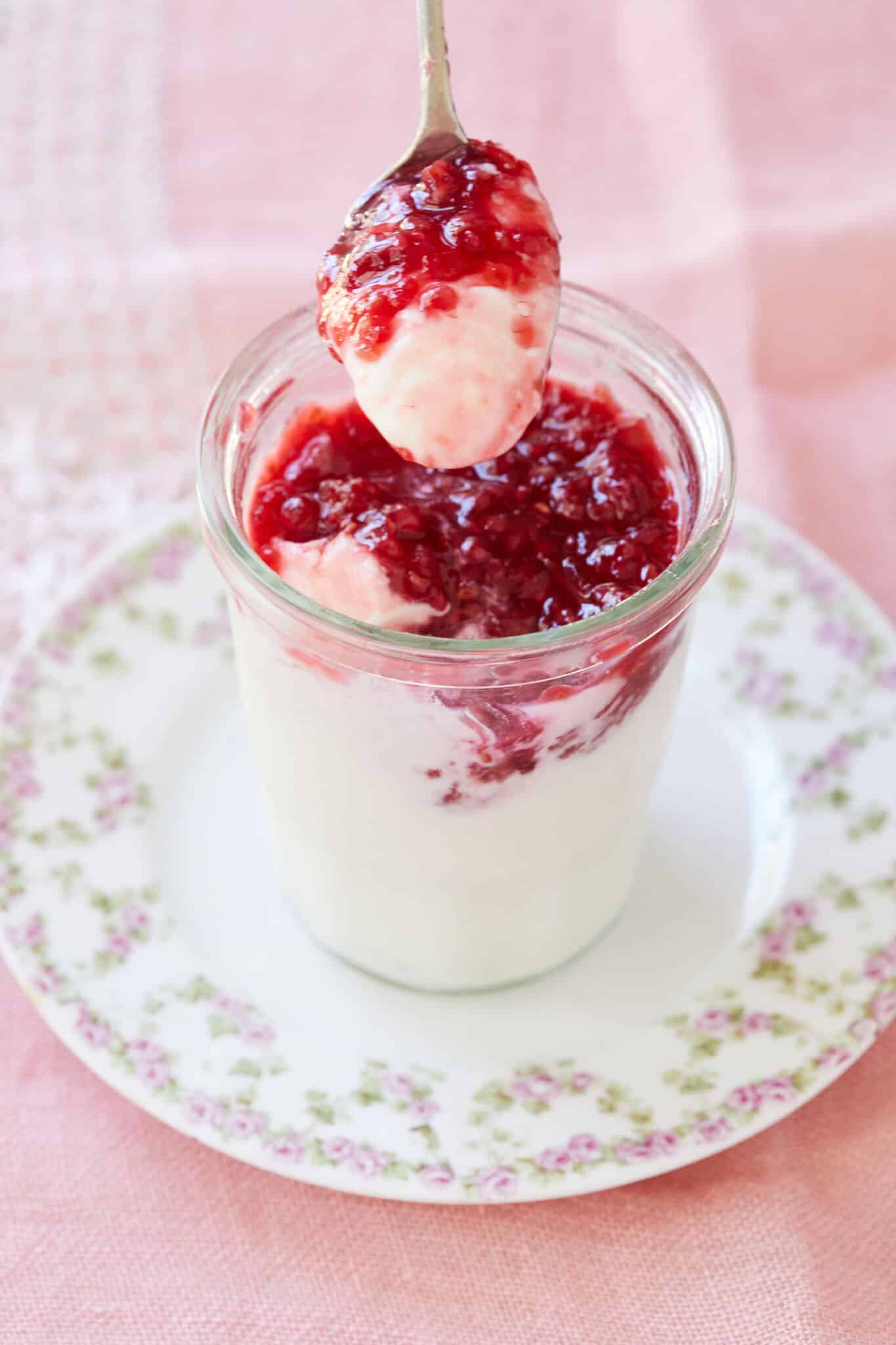 A spoonful of the white silky smooth buttermilk Panna Cotta with glossy vibrant red raspberry jam is being taken from the glass jar. It's served on a floral plate.