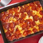A classic Detroit Style Pizza is baked in a rectangular metal pan, with thick and airy crust, crunchy caramelized edges, an ample amount of gooey and slightly toasted cheese and covered with crispy thin pepperoni.