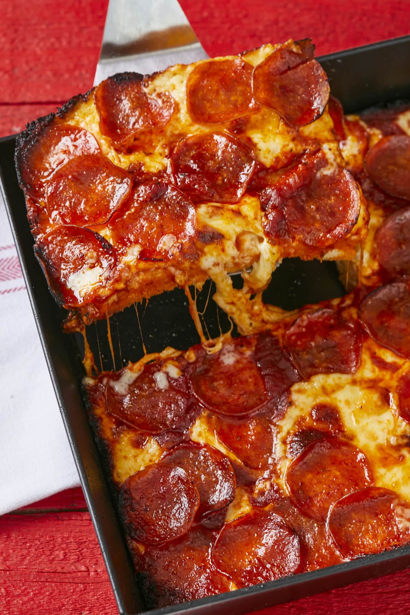 A slice of heavenly delicious pizza is cut and being lifted from the pan, with thick sand airy crust, crunchy caramelized edges, gooey toasted cheese, and thin crispy pepperoni. 