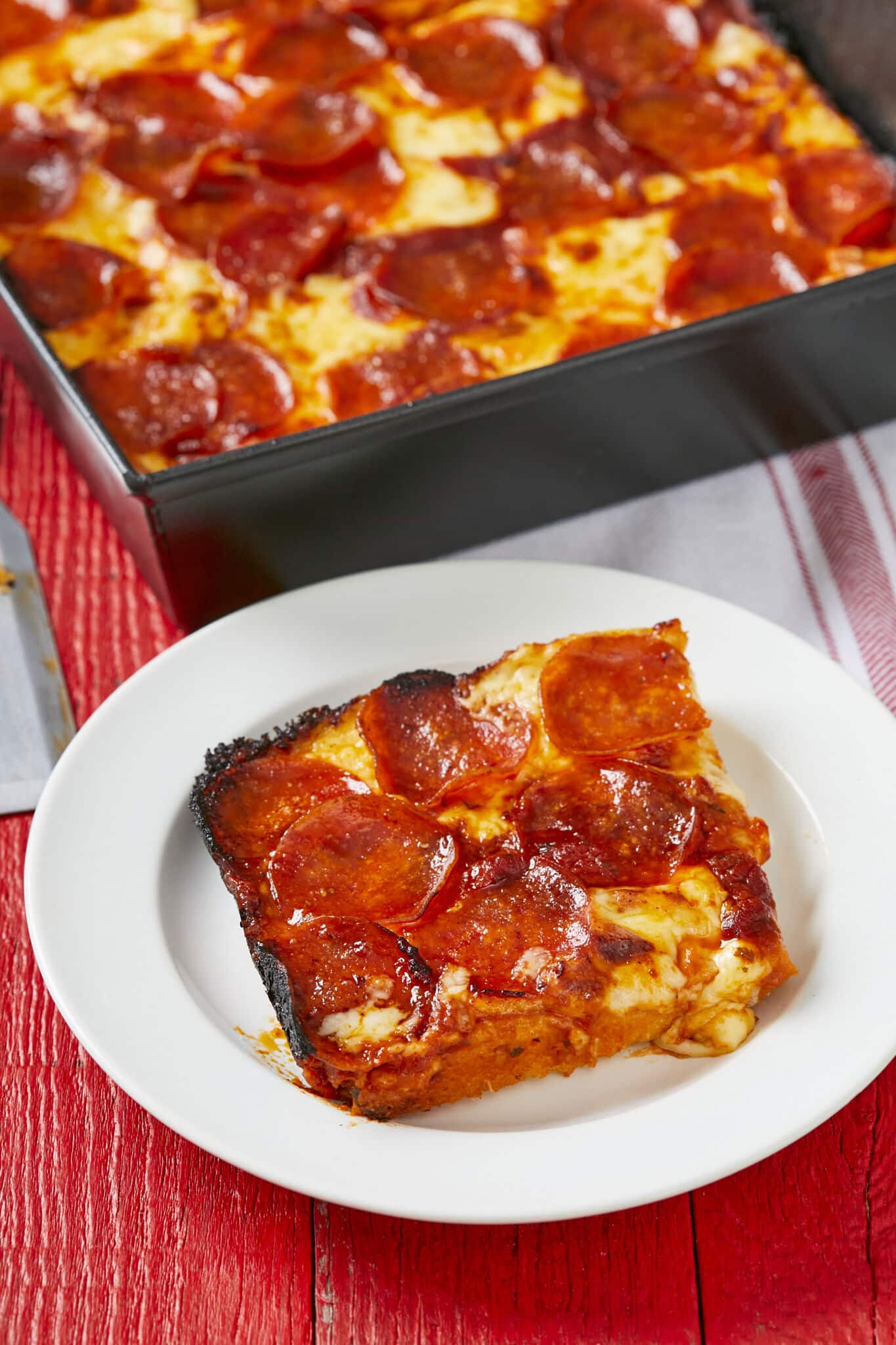 A slice of Detroit Style pizza is served on a white plat next to the remaining pizza in a black metal baking pan. It's featuring thick and airy crust, crunchy edges, gooey melted cheese and crispy pepperoni. 