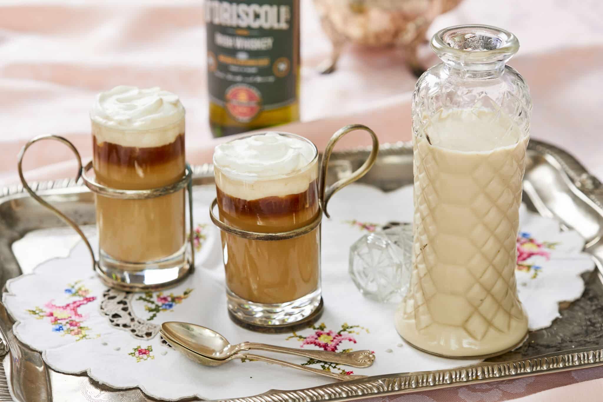 Silky smooth Homemade Irish Cream is in stored in a tall weave pattern glass bottle on the right side on a silver platter. To the left of the bottle are two glasses of Irish Cream Frappuccino which has blended sweet, floral, nutty and boozy Irish Cream with coffee and milk at the bottom, topped with pillowy whipped cream. A silver spoon is also on the platter next to the glasses. A bottle of O'Driscoll's whiskey and a tea pot are next to the platter in the upper center of the image.