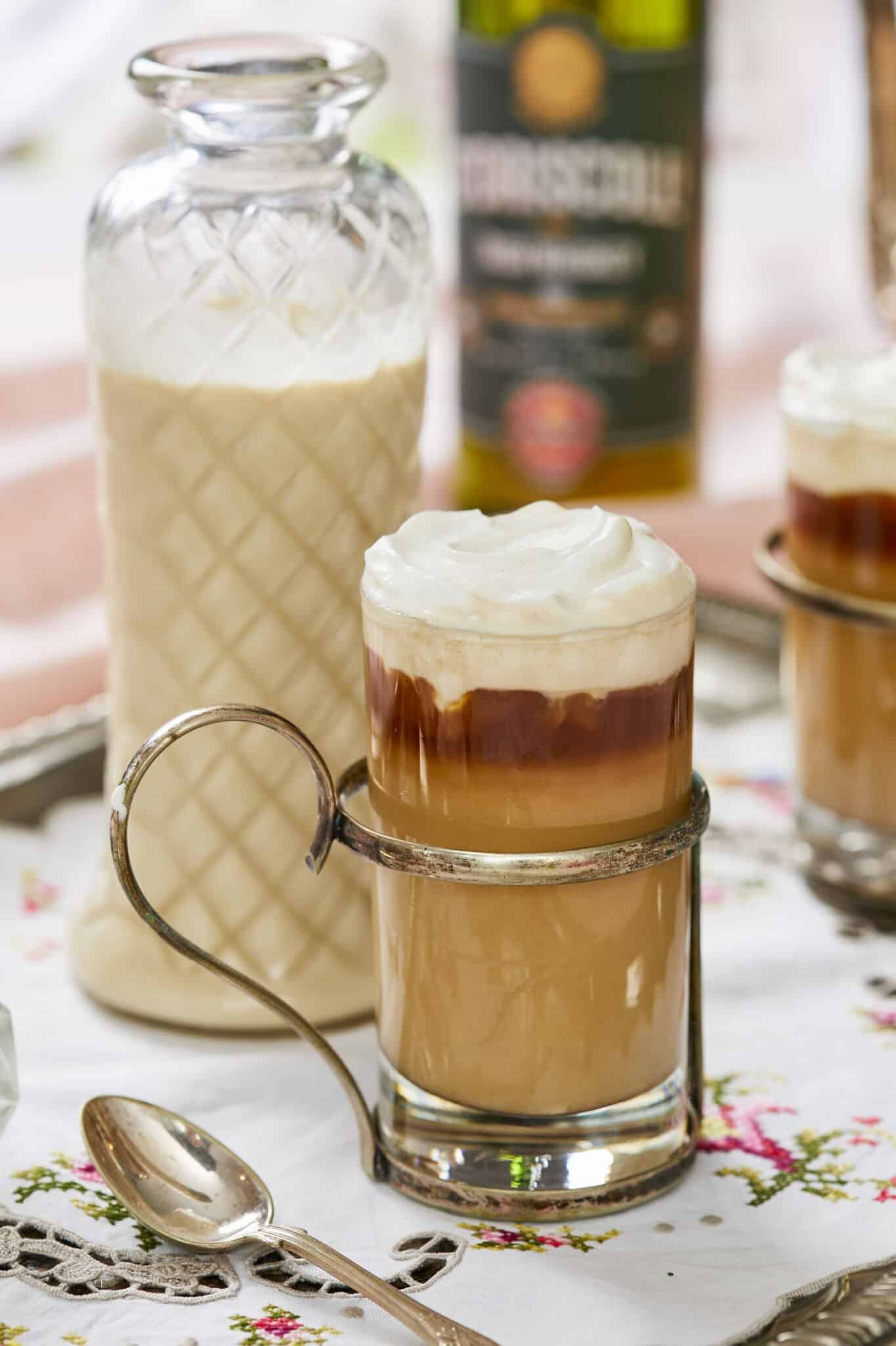 Silky smooth Homemade Irish Cream is in stored in a tall weave pattern glass bottle. In front of the bottle is a glass of Irish Cream Frappuccino which has layered blended sweet, floral, nutty and boozy Irish Cream with coffee and milk at the bottom, topped with pillowy whipped cream. A silver spoon is next to the glass. A bottle of O'Driscoll's whiskey and a second glass of frap are in the background. 
