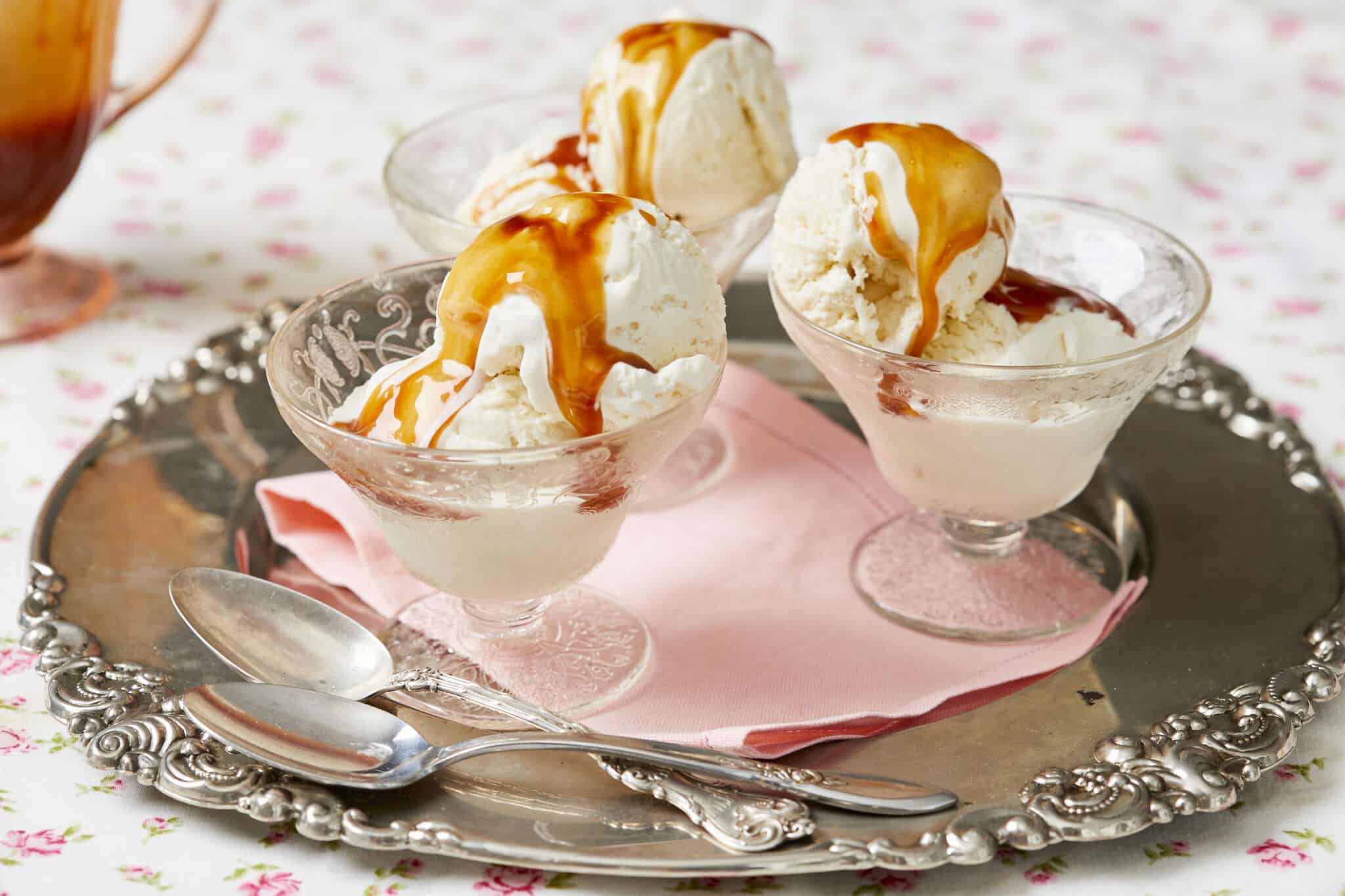 The velvety Irish Cream Ice Cream is served in 3 small glass dessert bowls on a pale pink napkin on a silver plater with floral patterns, drizzled with glossy dark-amber Irish whiskey caramel sauce.