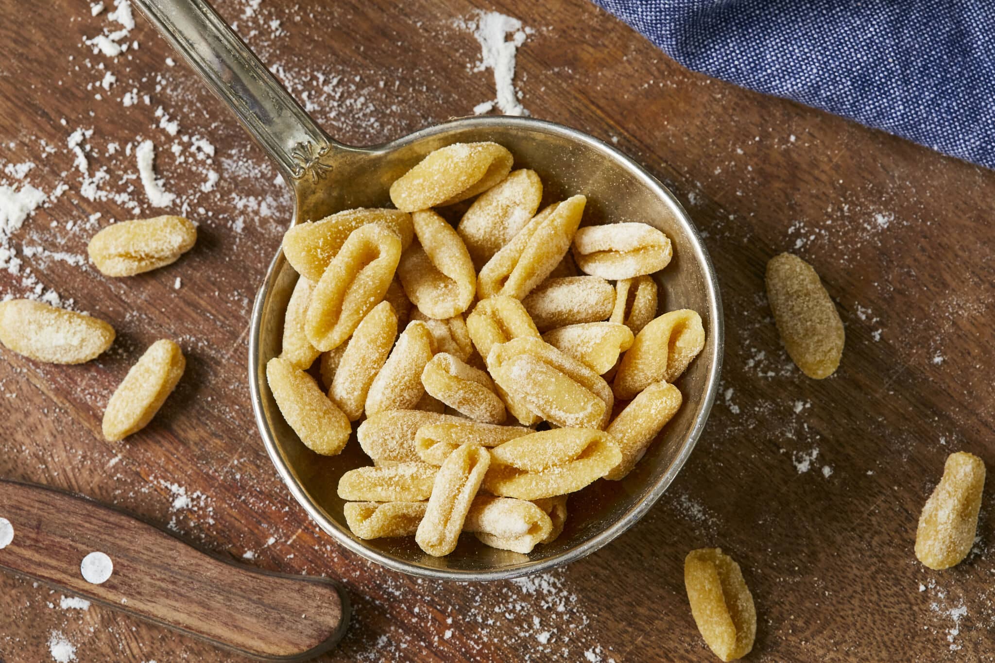 Cavatelli pasta is in a large metal ladle spoon. It has a curved shape of an elongated shell or a tiny hot dog bun! There are some extra pieces of cavtelli on the floured wooden board.