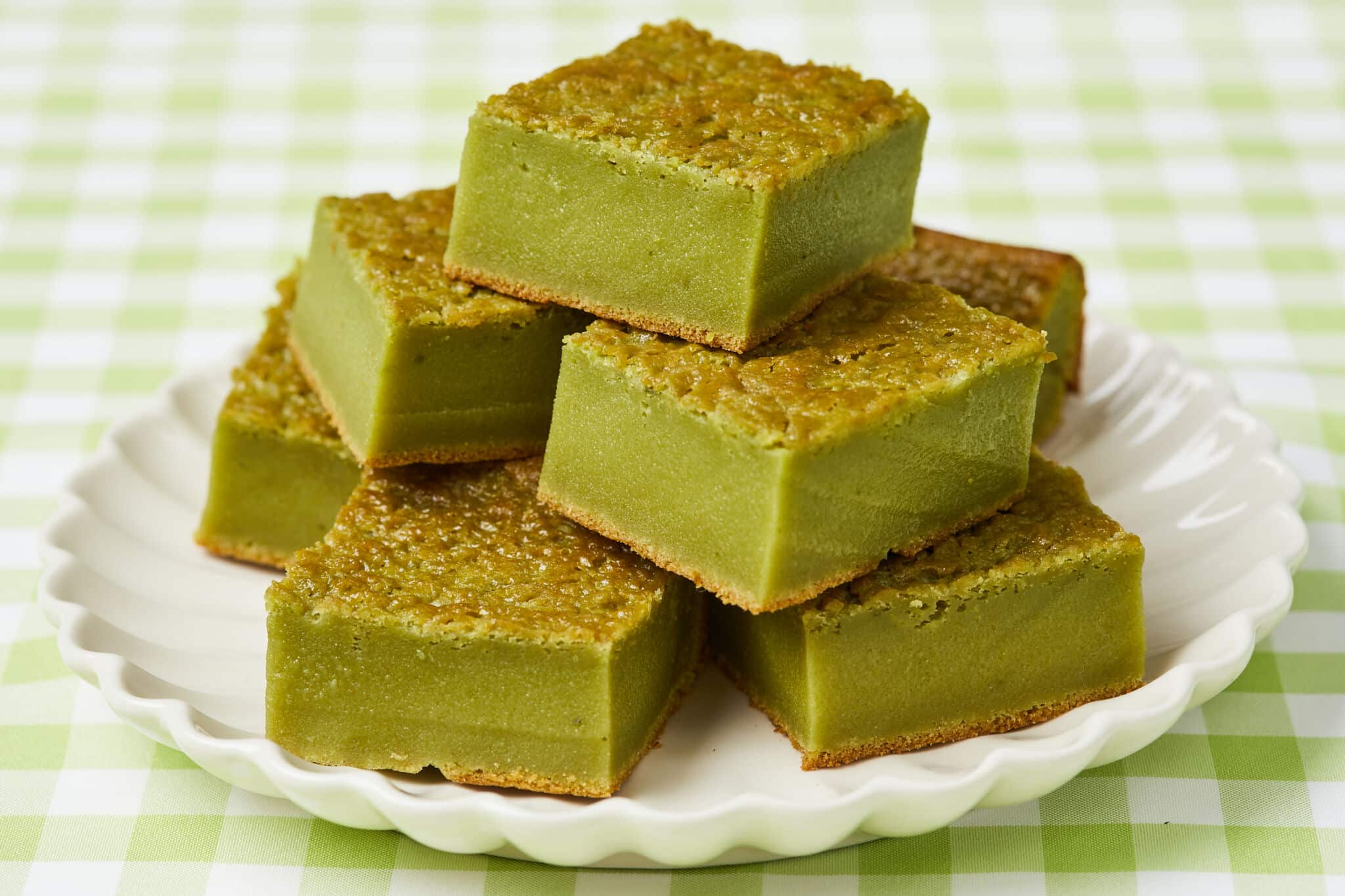 6 squares of rich Matcha Butter Mochi are placed on a white scalloped-edge plate. The green tea sweet rice cake has golden crispy crust on top and at the bottom, with green chewy and soft center. 