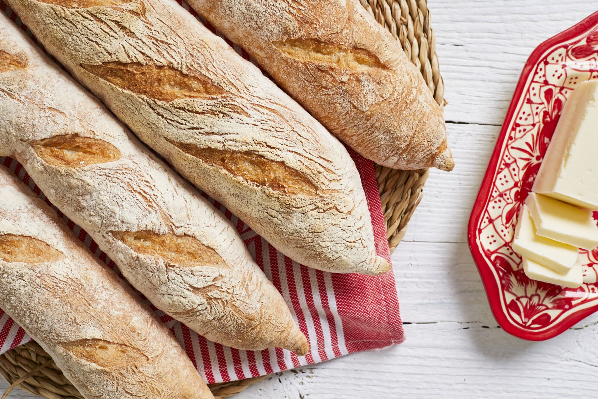 The Easiest No Knead Baguette Bread featuring a gorgeous thin, golden exterior are placed on a red-strip kitchen towel, paired with butter on a red butter dish.