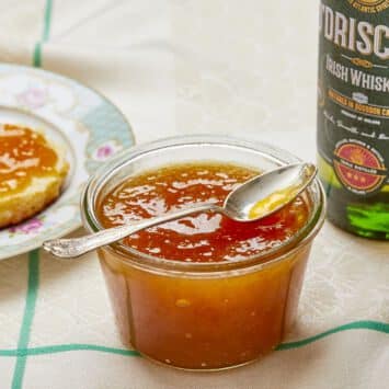 A glass jar of Sweet and Boozy Peach Whiskey Jam is in the middle in vibrant orange color and silky smooth texture. Half scone topped with this spread is on the top left, a bottle of O'Driscoll Irish whiskey is on the right.
