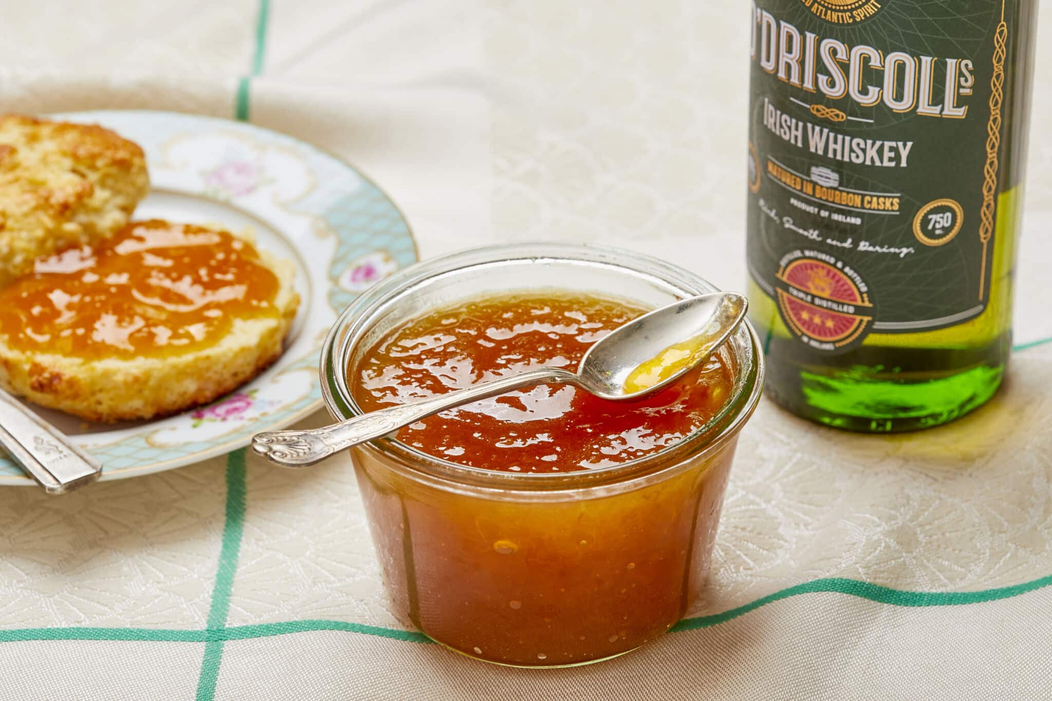 A glass jar of Sweet and Boozy Peach Whiskey Jam is in the middle in vibrant orange color and silky smooth texture. Half scone topped with this spread is on the top left, a bottle of O'Driscoll Irish whiskey is on the right.