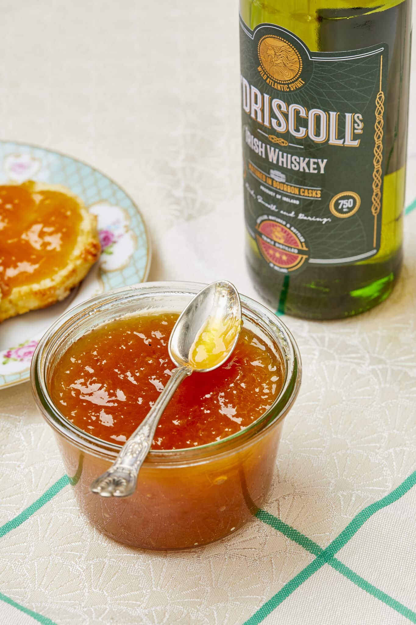 A glass jar of Sweet and Boozy Peach Whiskey Jam is in the middle in vibrant orange color and silky smooth texture. Half scone topped with this spread is on the top left, a bottle of O'Driscoll Irish whiskey is on the right. 