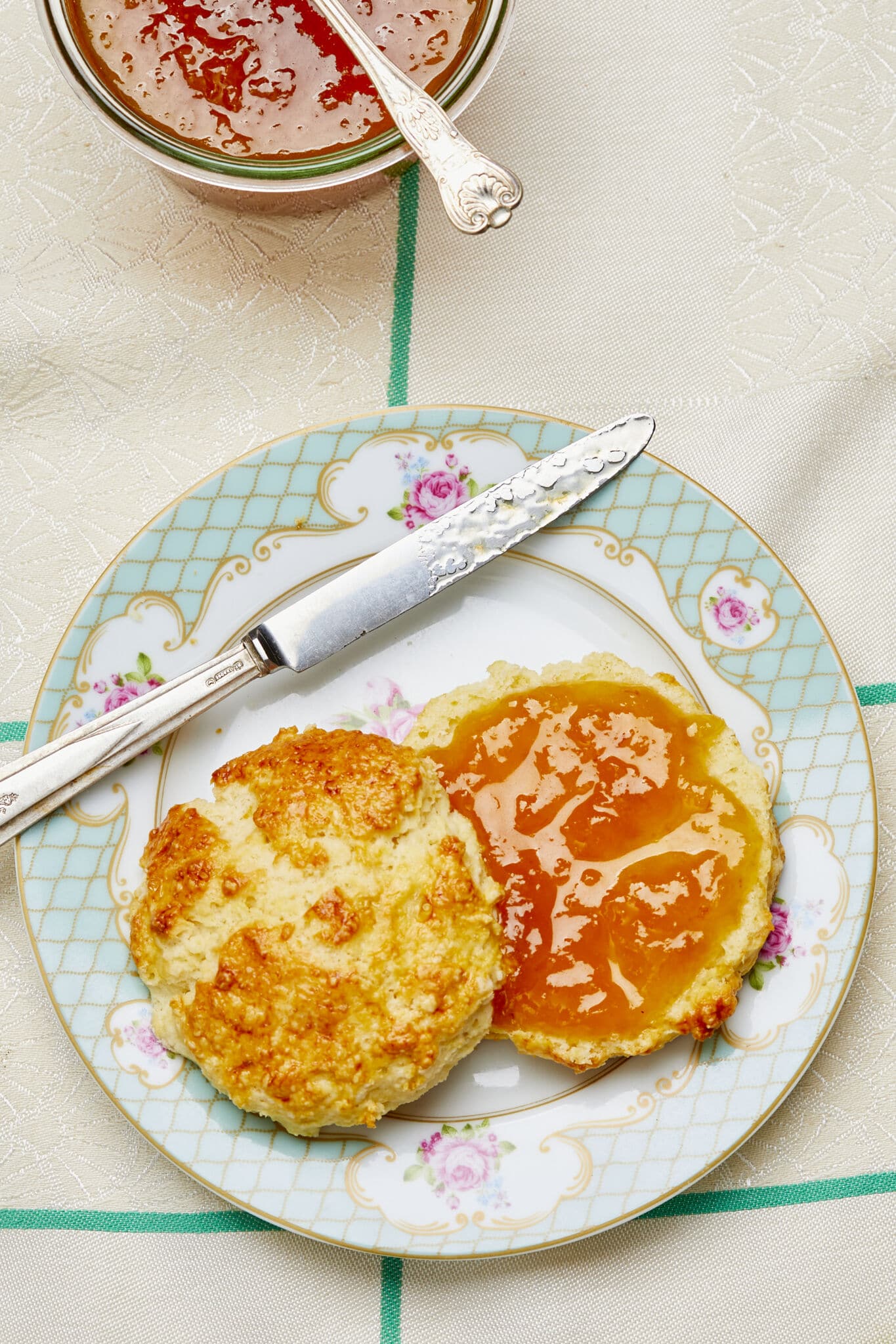 A scone with golden crust is cut in half, the bottom piece is smeared with velvety jam in bright orange color. A jar of Peach Whiskey jam is served on the side with a spoon. 