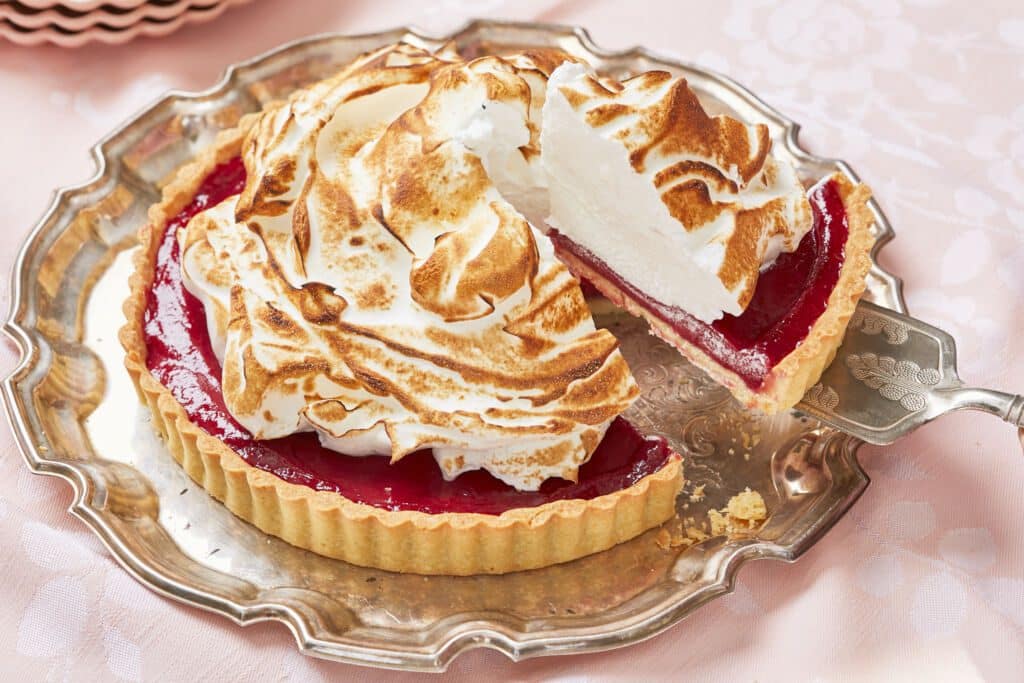 A slice of the Mile-High Raspberry Meringue pie is cut from a silver pate. The side shows its buttery rich pie crust filled with vibrant red fruity raspberry filling, topped with a generous amount of toasted golden brown and light glossy meringue.