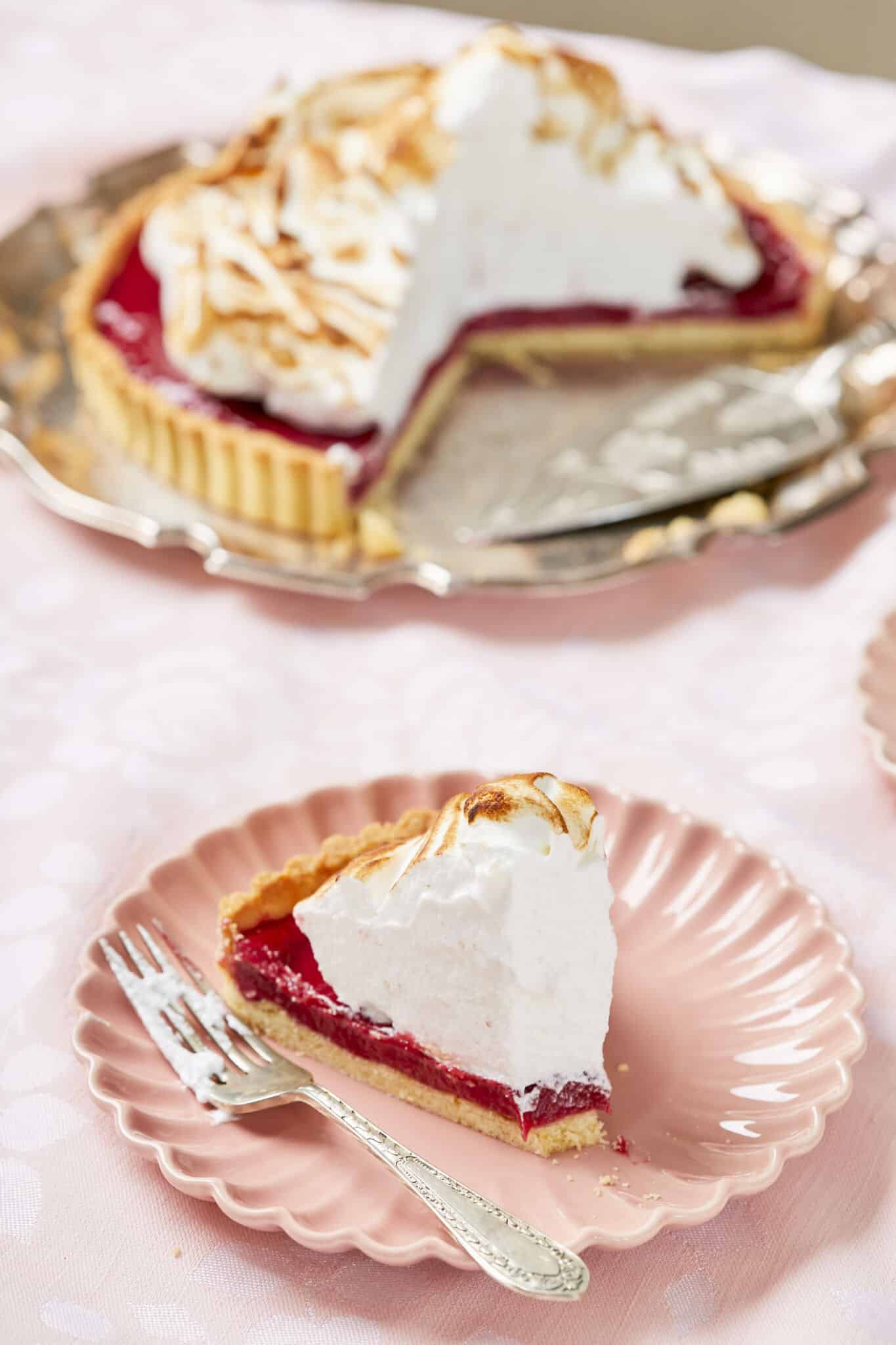 A slice of the pie is served on a small dessert plate next to the main pie. One bite is taken from the small slice, showing the buttery crust, vibrant read tangy raspberry curd topped with a generous amount of fluffy, golden and crispy meringue. 
