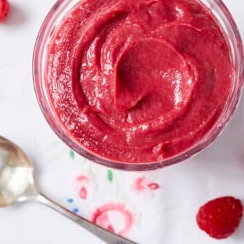 An overhead shot of a glass bowl of stunning and luscious raspberry curd in vibrant red color and super creamy texture. A spoon and three fresh raspberries are on the white linen with embroidered flowers outside the bowl.