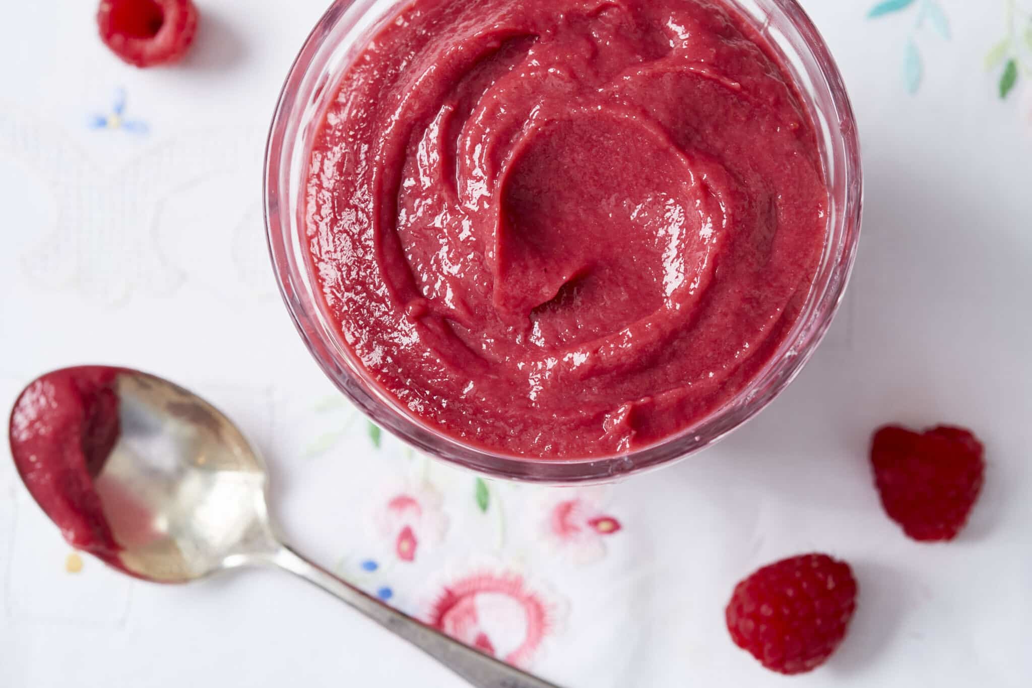 An overhead shot of a glass bowl of stunning and luscious raspberry curd in vibrant red color and super creamy texture. A spoon and three fresh raspberries are on the white linen with embroidered flowers outside the bowl.