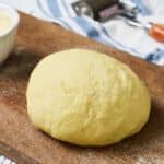 Semolina pasta dough is resting after kneading on a wooden board. It's a golden-hued mixture with a slightly coarse texture. When combined, it forms a pliable and firm mass that is smooth to the touch. The dough feels substantial yet manageable, allowing for easy shaping and rolling.