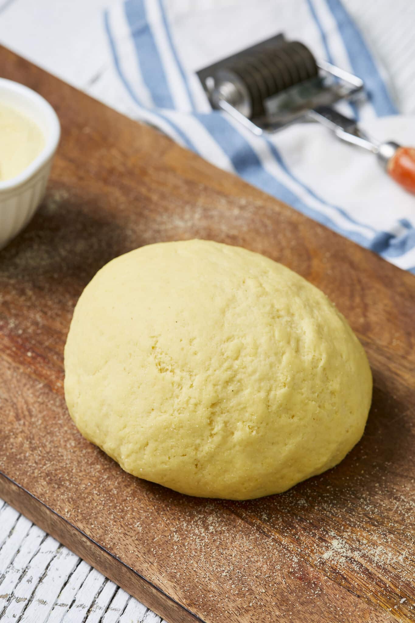 A yellow, smooth, and round dough is resting on a wooden board before using. A pasta roller is on the side. 