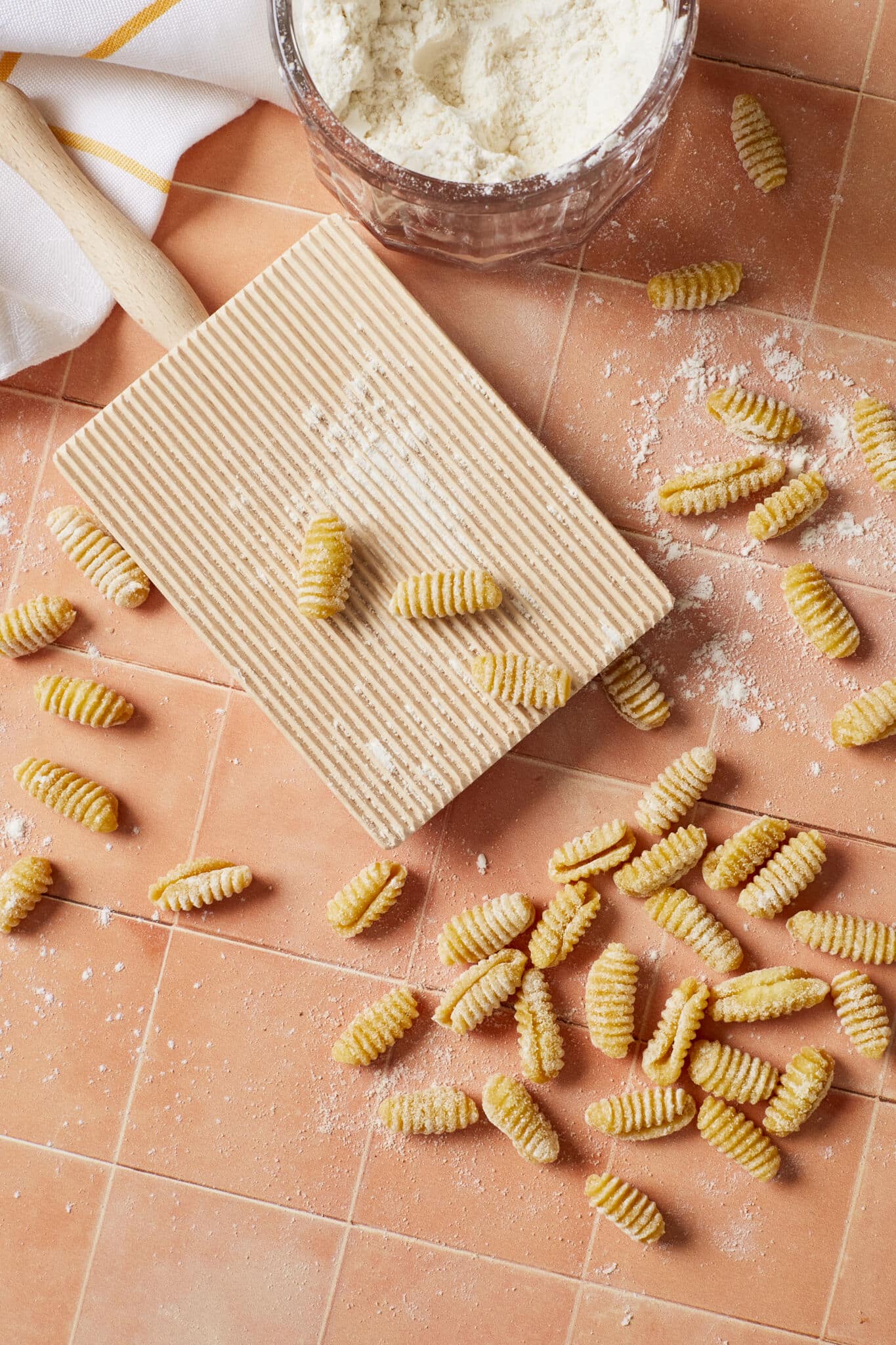 Malloreddus Pasta (Gnocchetti Sardi) is placed around a gnocchi board with 3 pieces on it. Malloreddus is shaped into small, elongated shells with a slightly curved shape. A glass bowl of flour is on the side. 