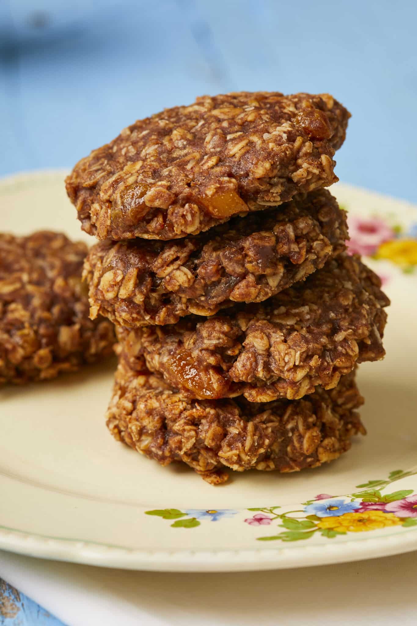 A stack of four breakfast cookies are placed on a plate with one in the back. They're thick and golden brown, packed with delicious and healthy rolled oats, dried fruit, coconut flakes, mashed banana, eggs, peanut butter and honey. 