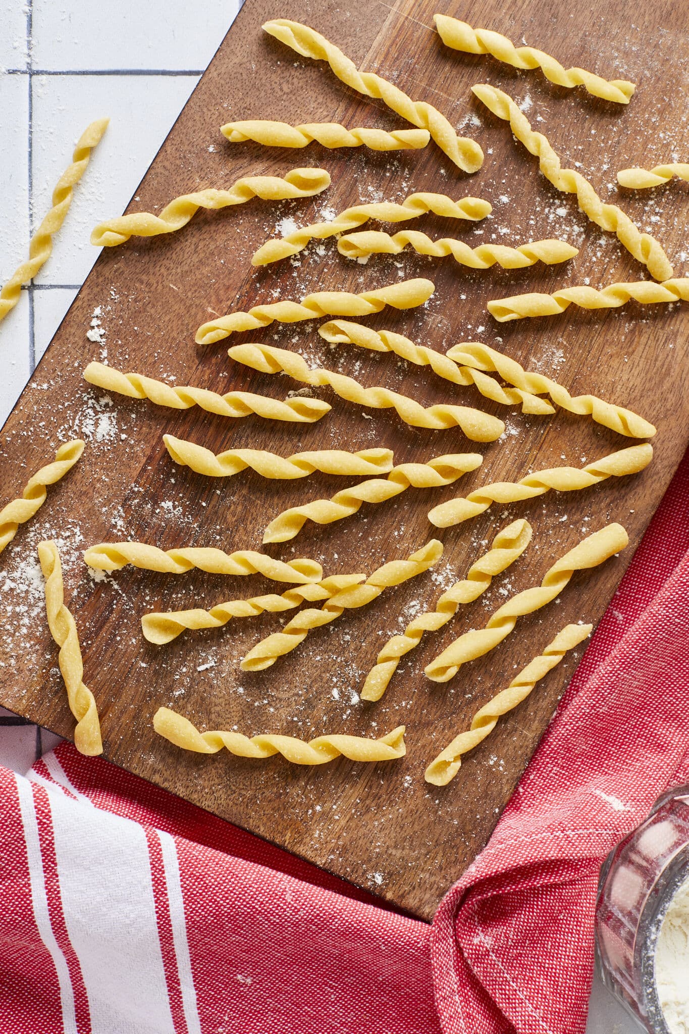 Yellow semolina Busiate pasta is in characteristic cord shape and placed on a lightly floured wooden board, with a red color kitchen towel underneath. 