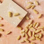 Malloreddus Pasta (Gnocchetti Sardi) is placed around a gnocchi board with 3 pieces on it. Malloreddus is shaped into small, elongated shells with a slightly curved shape. A glass bowl of flour is on the side.