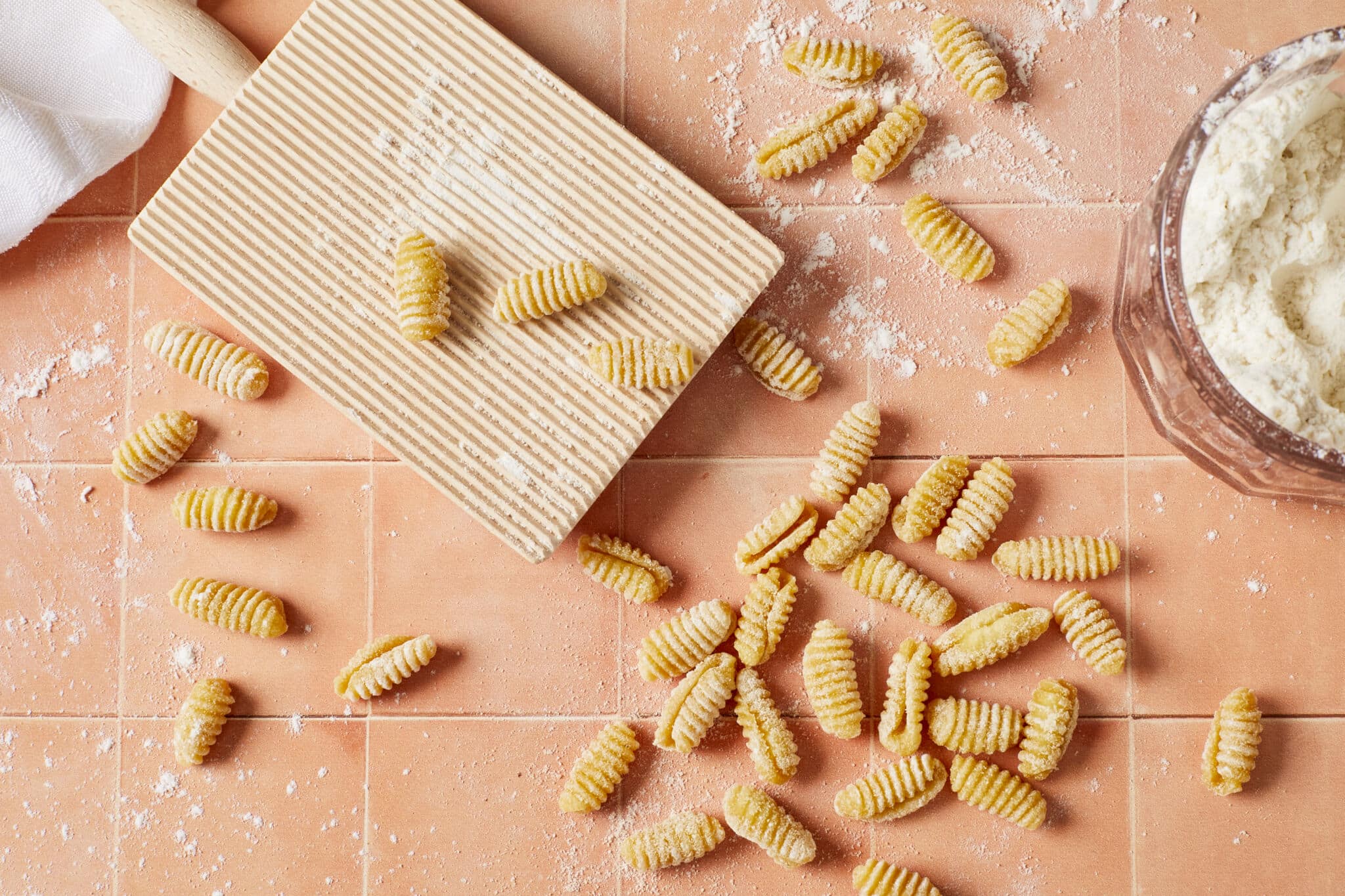 Malloreddus Pasta (Gnocchetti Sardi) is placed around a gnocchi board with 3 pieces on it. Malloreddus is shaped into small, elongated shells with a slightly curved shape. A glass bowl of flour is on the side.