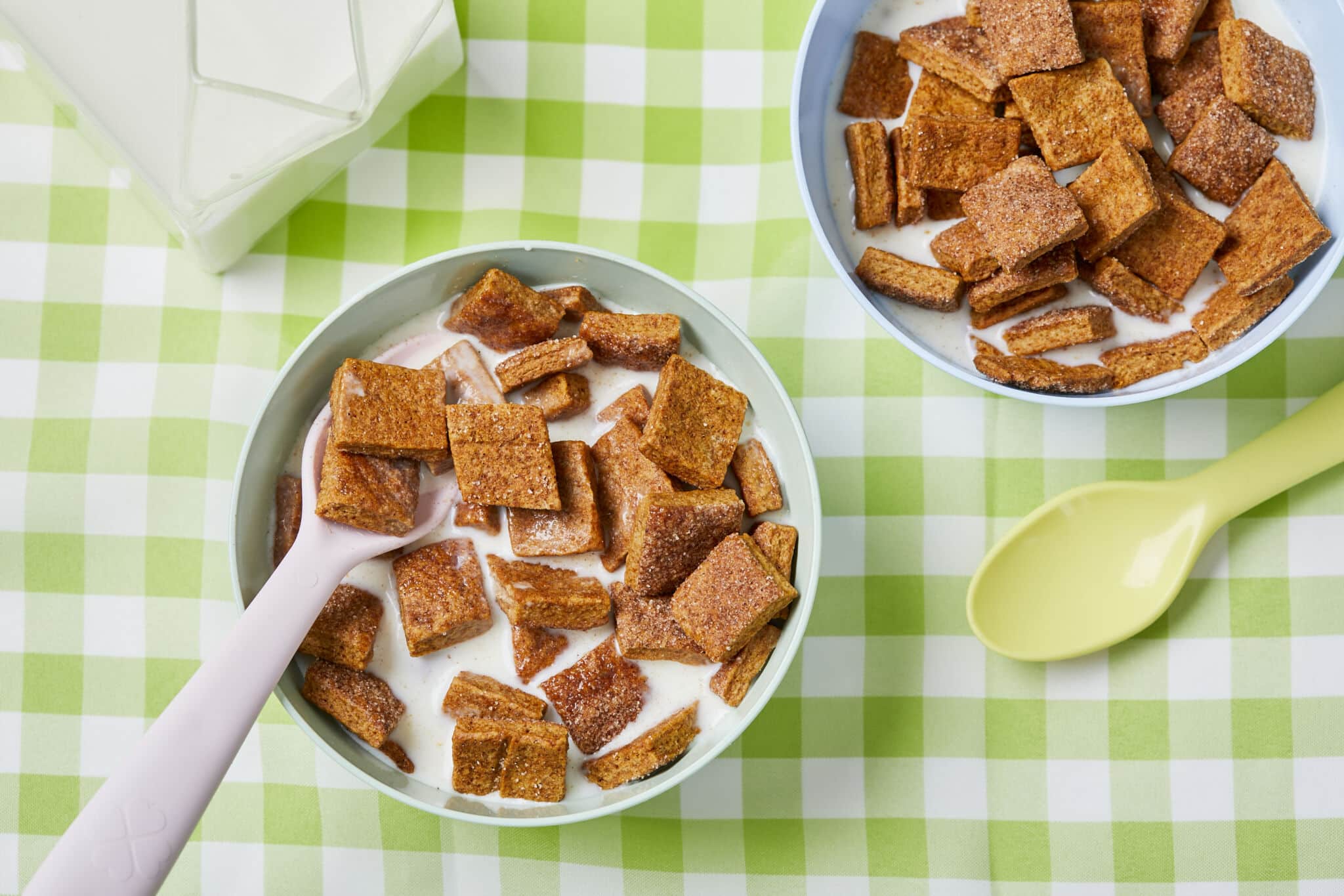 Two bowls of Homemade Cinnamon Toast Crunch cereal is served with milk and spoons with the milk bottle on the left side. The cereal has hearty, extra-crispy squares of sweet, warmly-spicy cinnamon flavor. This wholesome version is a welcome and cozy option for breakfast or a snack.
