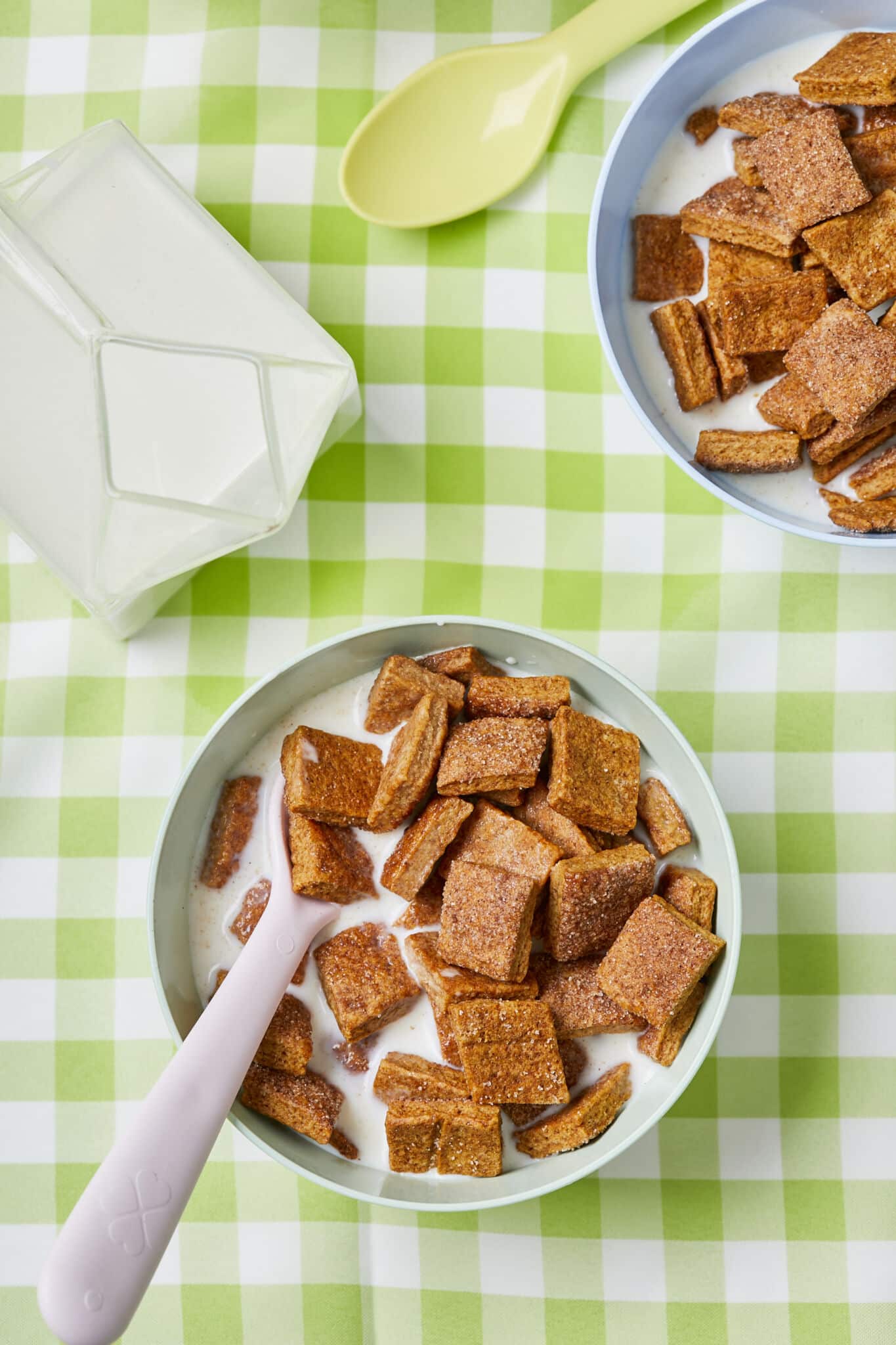 Two bowls of Homemade Cinnamon Toast Crunch cereal is served with milk and spoons with the milk bottle on the left side. The cereal has hearty, extra-crispy squares of sweet, warmly-spicy cinnamon flavor. This wholesome version is a welcome and cozy option for breakfast or a snack. 