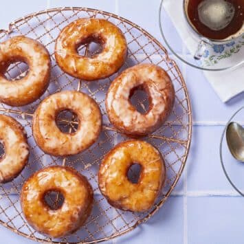 Homemade Krispy Kreme donuts are placed on a round rose-color wire cooling rack. These donuts are fried perfectly golden brown and soft, and topped with glossy clear icing. Two cups of tea is served on the side.