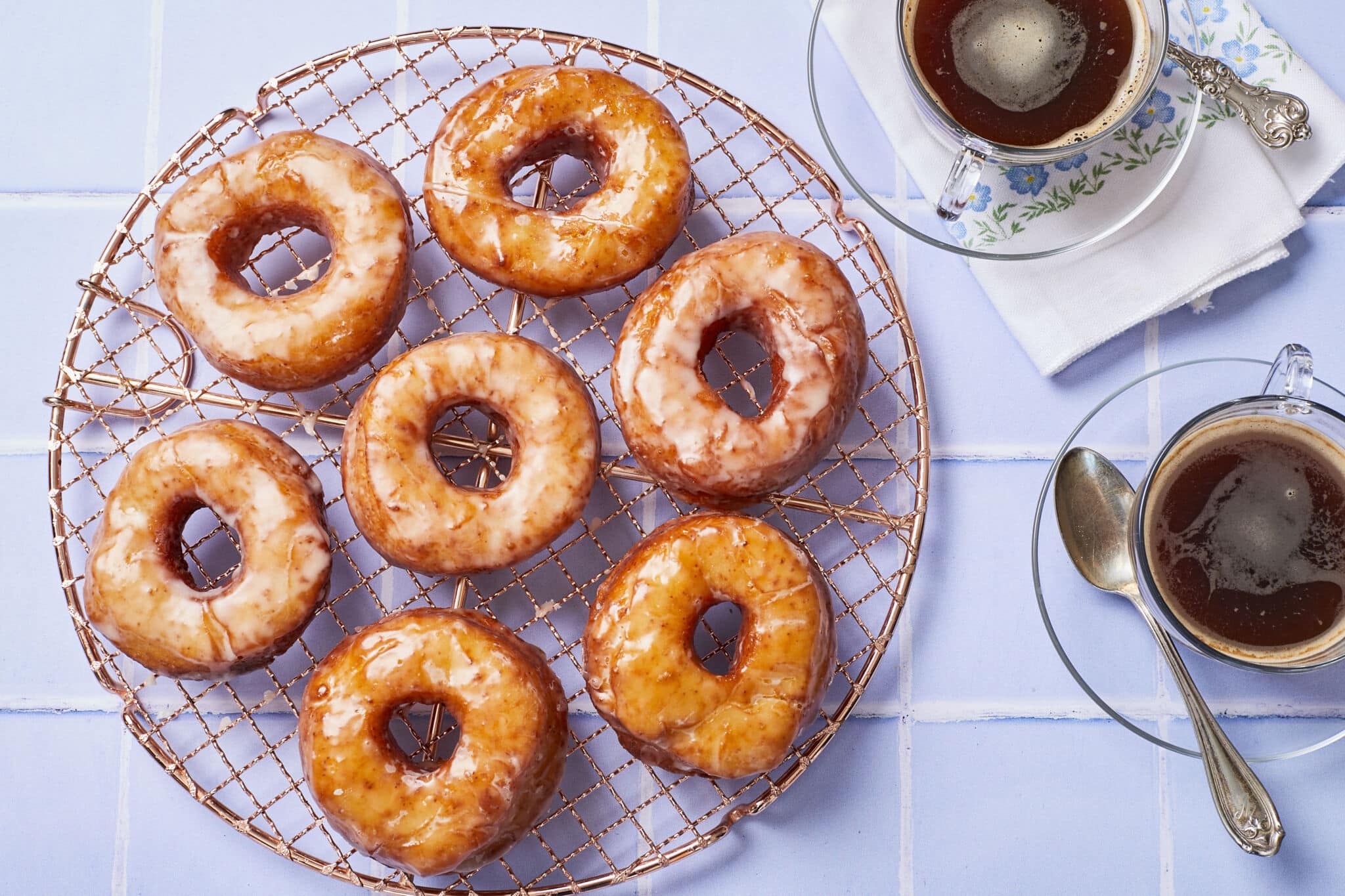 Homemade Krispy Kreme donuts are placed on a round rose-color wire cooling rack. These donuts are fried perfectly golden brown and soft, and topped with glossy clear icing. Two cups of tea is served on the side.