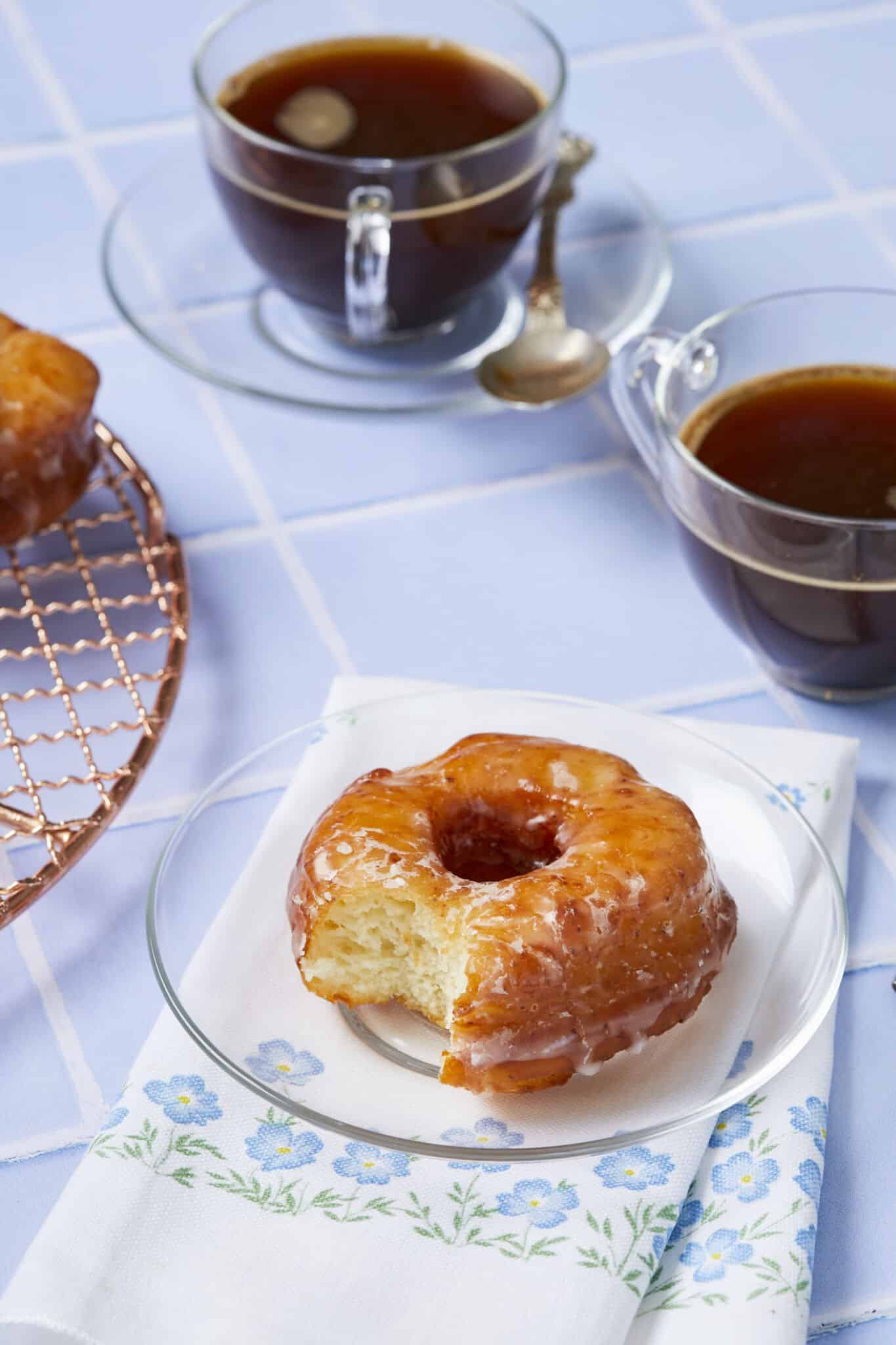 A close shot at a perfectly fried and glazed donut on a clear glass dessert plate, with one bite taken. The interior looks light with lots of air pockets. Served with tea. 