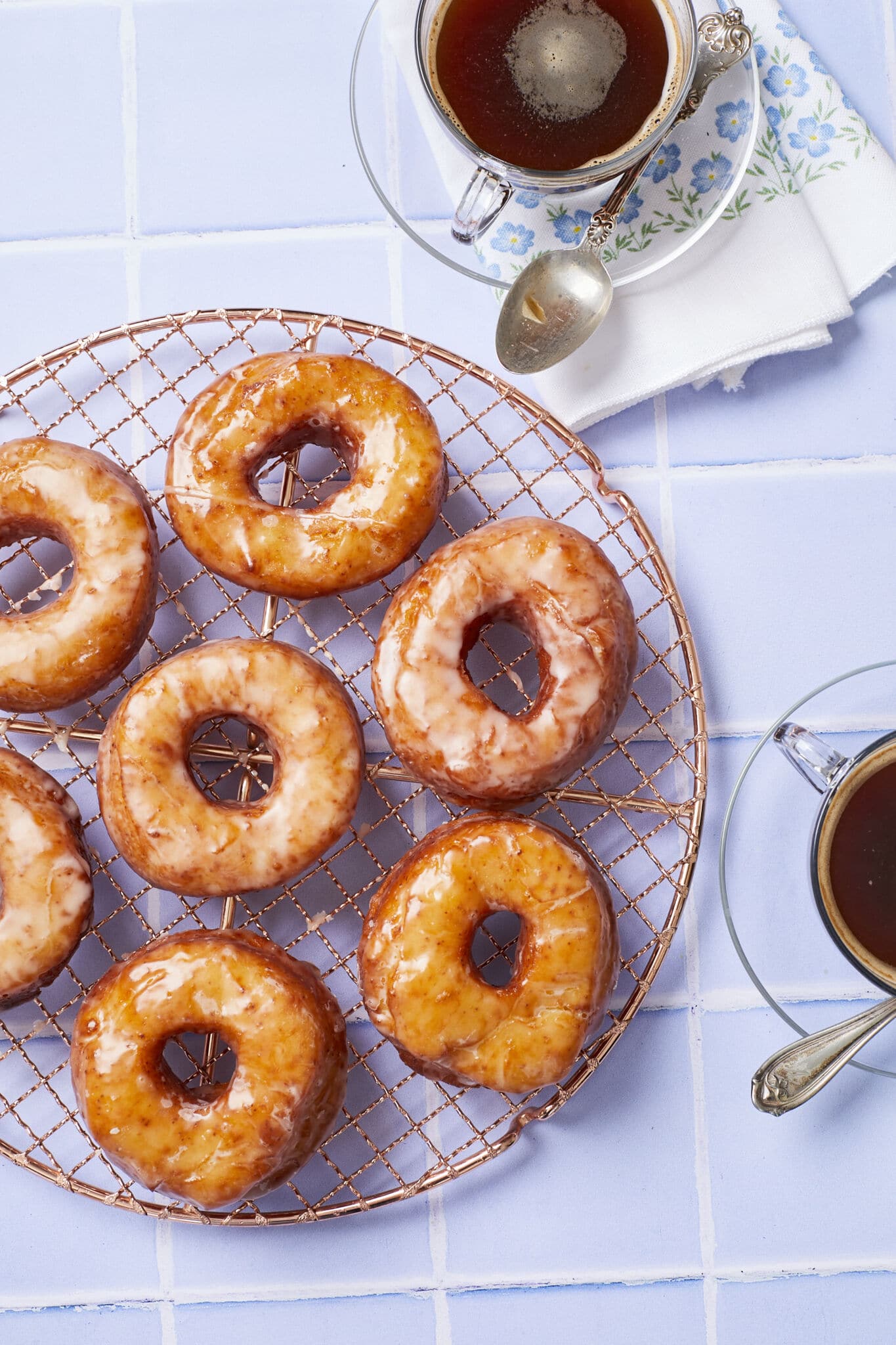 Homemade Krispy Kreme donuts are placed on a round rose-color wire cooling rack. These donuts are fried perfectly golden brown and soft, and topped with glossy clear icing. Two cups of tea is served on the side. 