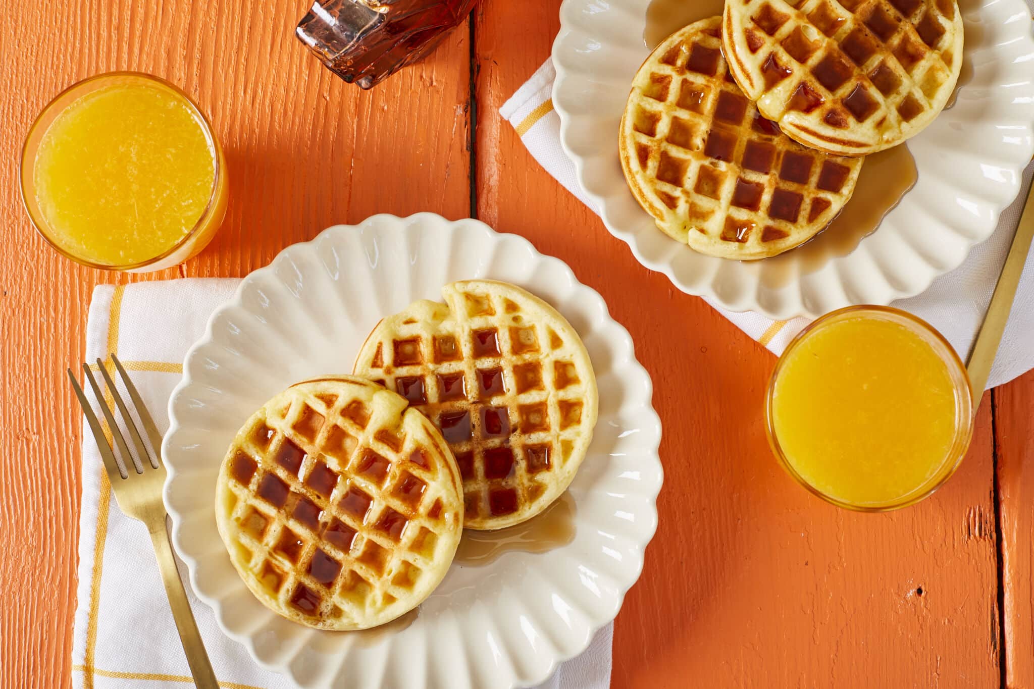 Four Homemade Eggo waffles are served with two on each white plate. The waffles are golden with crispy edges and tender interior, drizzled with maple syrup and paired with a glass of orange juice on the side.