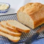 5-Ingredient No-Knead Sandwich Bread is sliced and cooling on a black wire rack with a bread knife next to it. The bread has a golden crisp crust and a soft, tender even crumb. Butter is served on the side to pair with the bread.