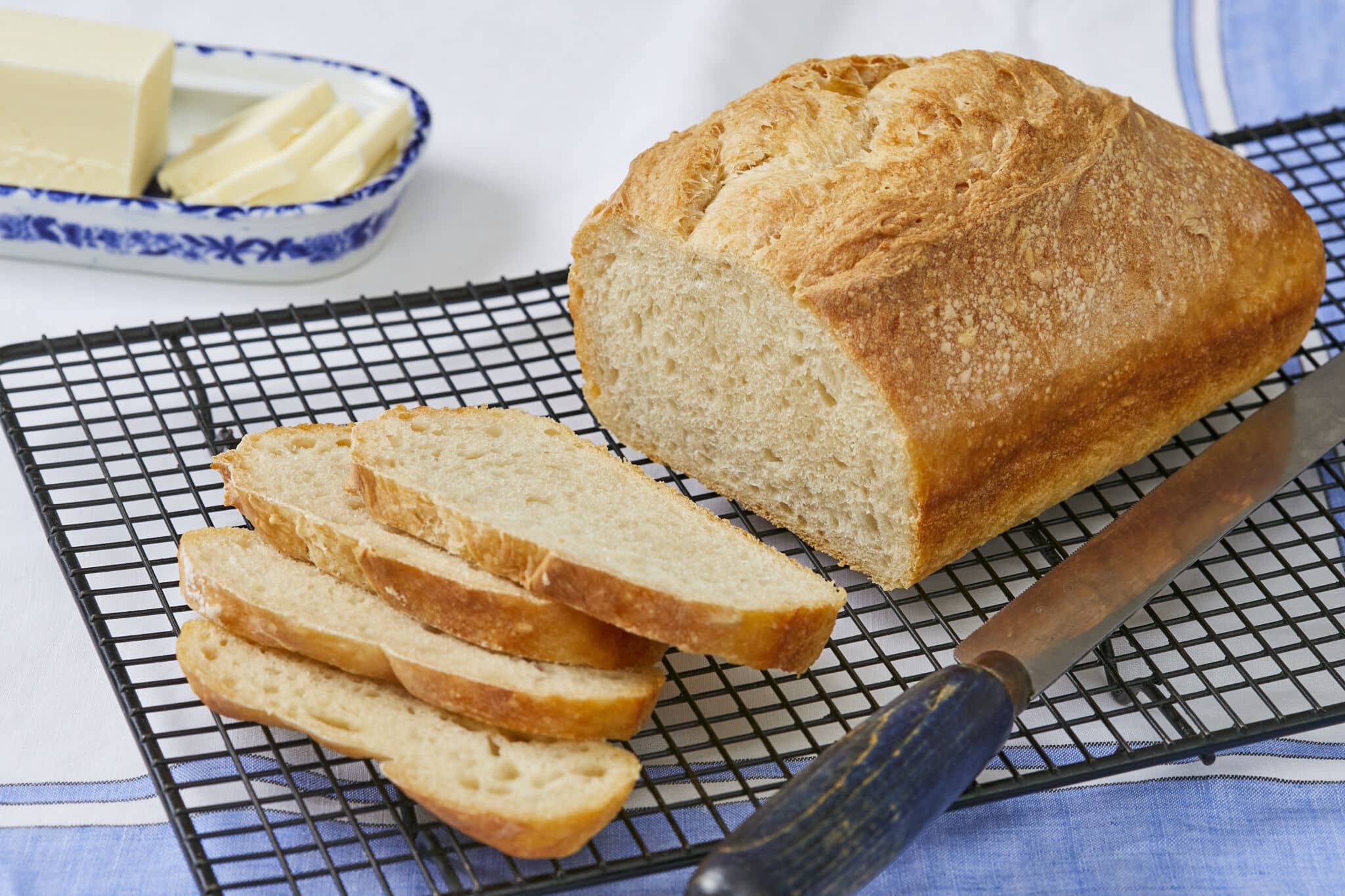 5-Ingredient No-Knead Sandwich Bread is sliced and cooling on a black wire rack with a bread knife next to it. The bread has a golden crisp crust and a soft, tender even crumb. Butter is served on the side to pair with the bread.
