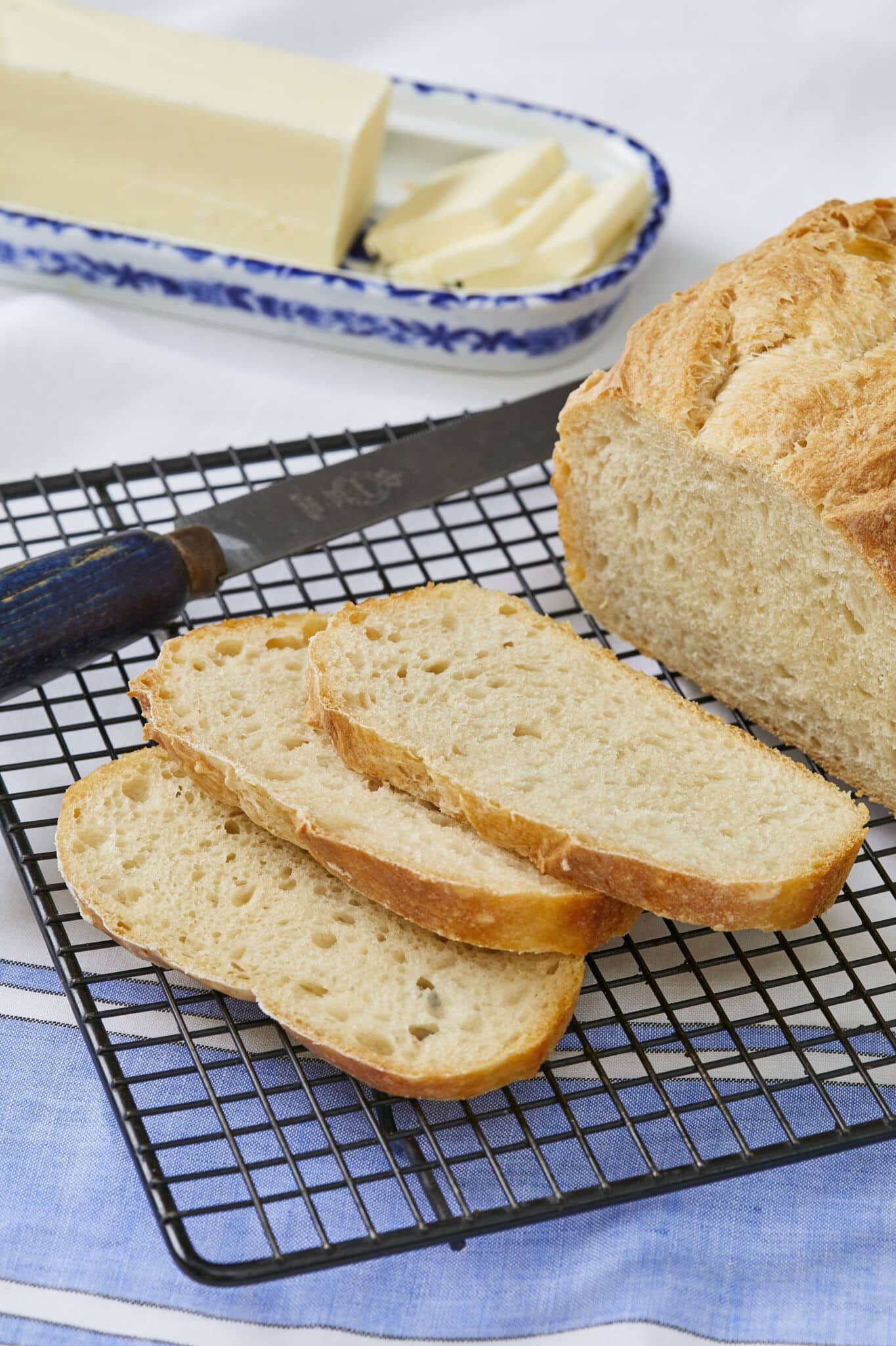 5-Ingredient No-Knead Sandwich Bread is sliced and cooling on a black wire rack with a bread knife next to it. The bread has a golden crisp crust and a soft, tender even crumb. Butter is served on the side to pair with the bread. 