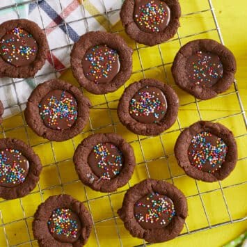 Bite-size Chocolate Cookie Cups are placed on a cooling rack. They are in signature chocolate brown color, have lightly crispy edges, soft and chewy inside with a silky ganache filling. They're decorated with colorful sprinkles. A glass cup of milk is served on the side.