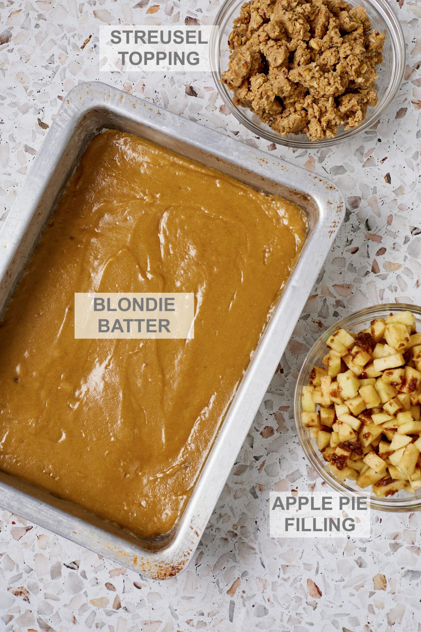 Key ingredients for Apple Pie Blondies include blondie batter in a metal baking tray, streusel topping in a glass bowl on the upper right side of the image, apple pie filling with chunks of apple and cinnamon sugar near the bottom right side of the image. 