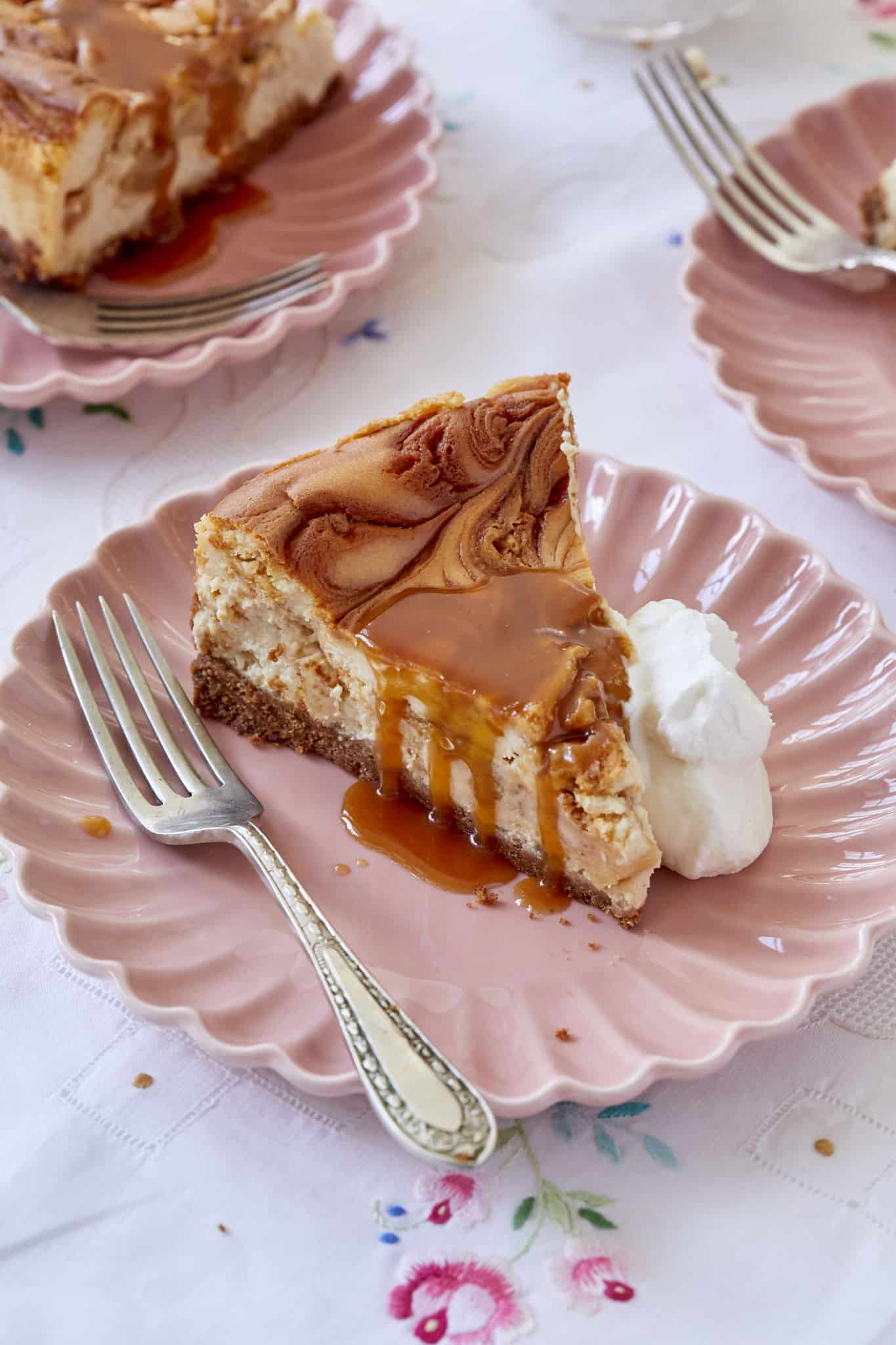 Decadent Caramel Apple Cheesecake is served on pink scallop-edged plates. It has buttery, crumbly graham cracker crust at the bottom, silly rich filling in the middle with a swirly top. It's paired with caramel sauce and a dollop of whipped cream. 