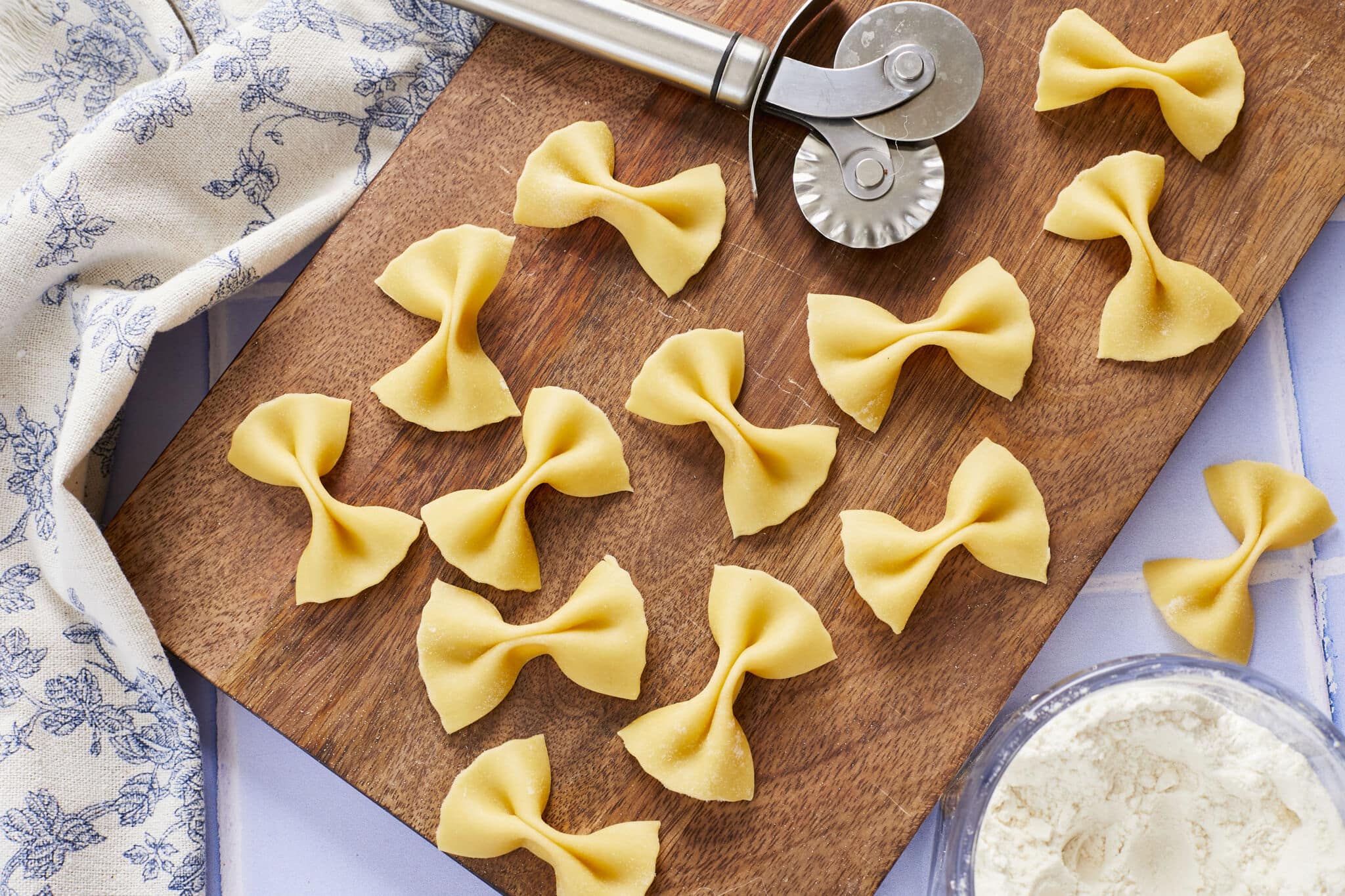 Butterfly-shaped Farfalle Pasta with perfect crevasses and fluted edges is drying on a wooden board with a fluted pasta cutter. A bowl of extra flour is on the side.