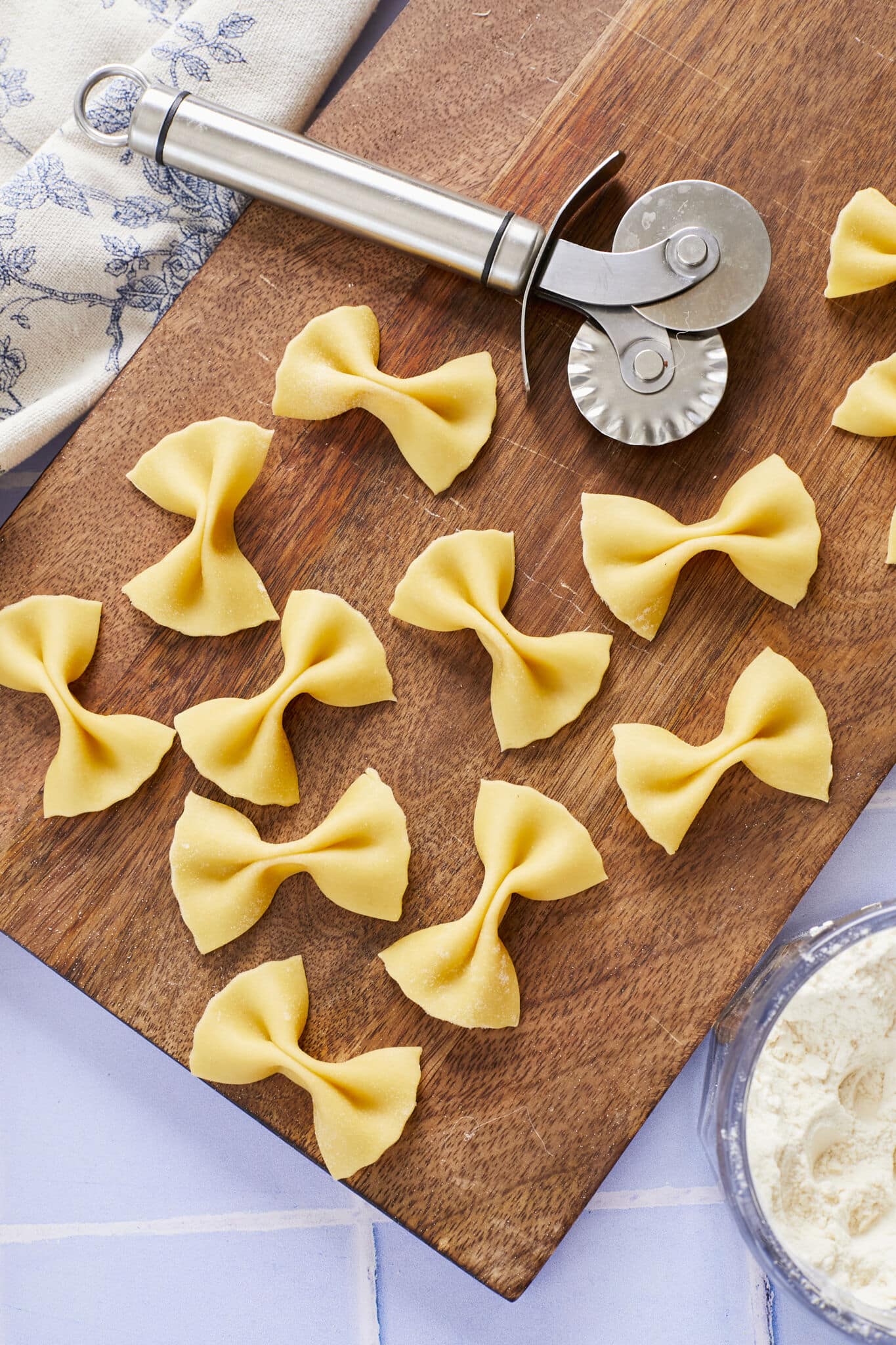 Butterfly-shaped Farfalle Pasta with perfect crevasses and fluted edges is drying on a wooden board with a fluted pasta cutter. A bowl of extra flour is on the side. 