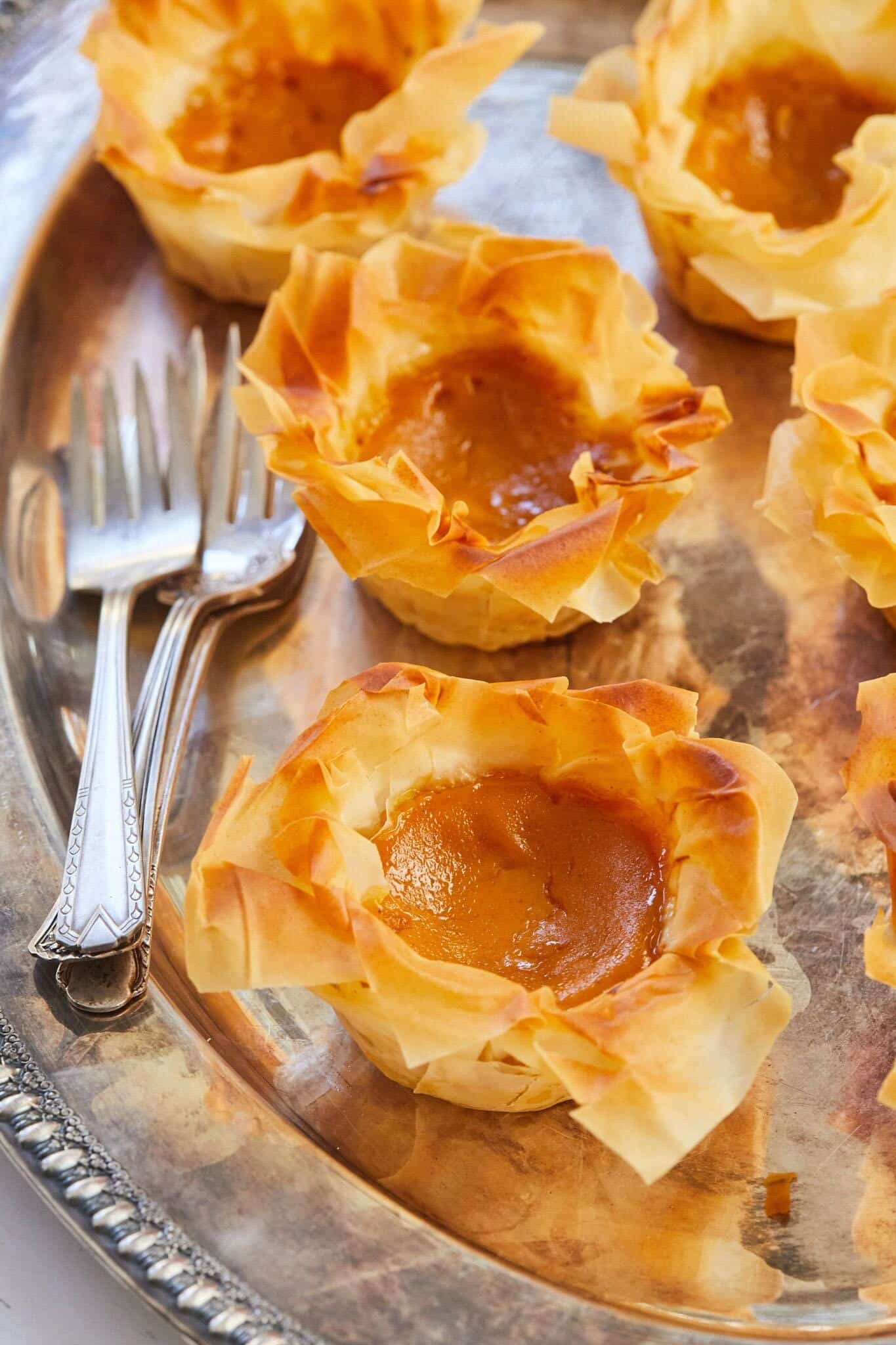 Petite Pumpkin Pies look gorgeous with golden crispy phyllo crust and golden orange silky smooth filling. They're served on a big silver pater with forks. 