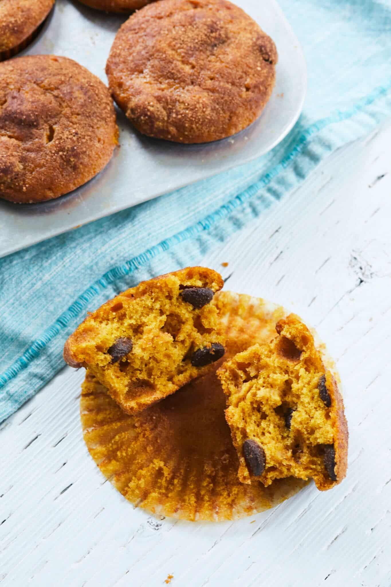 Chocolate Chip Pumpkin Muffins are baked to a perfect golden orange color with sprinkled cinnamon sugar on top. A muffin is cut into halves, showing the soft and moist inside and loaded with chocolate chips.