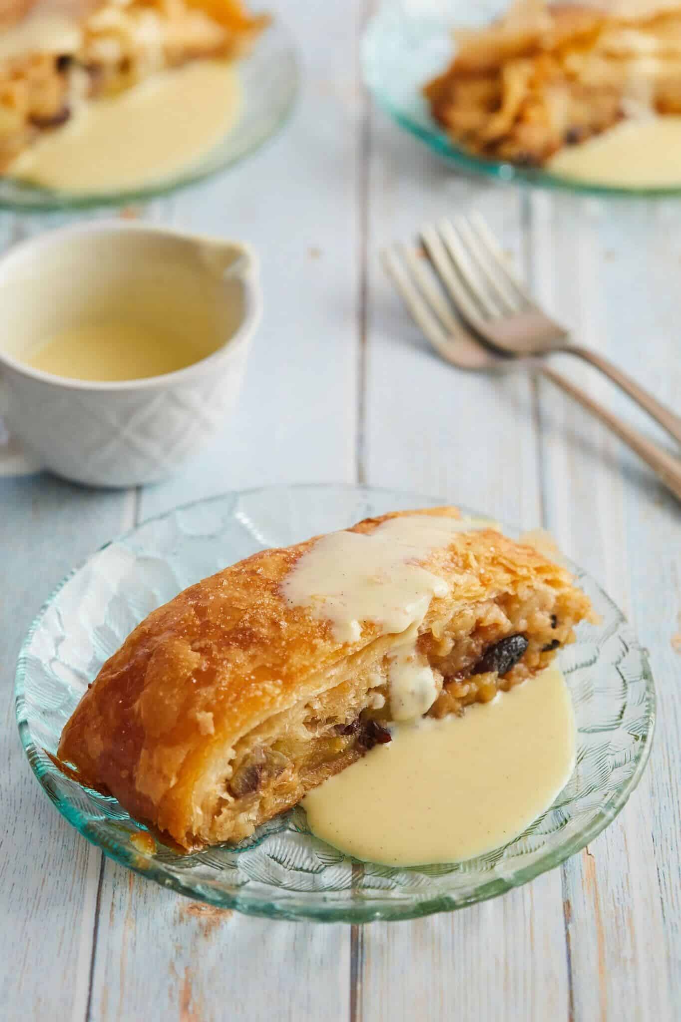 Apple Strudel is served on a glass plate covered in Creme Anglaise. The tender apples, aromatic cinnamon sugar are wrapped with layers of flaky pastry . More slices and Creme Anglaise are served on the side with two forks.