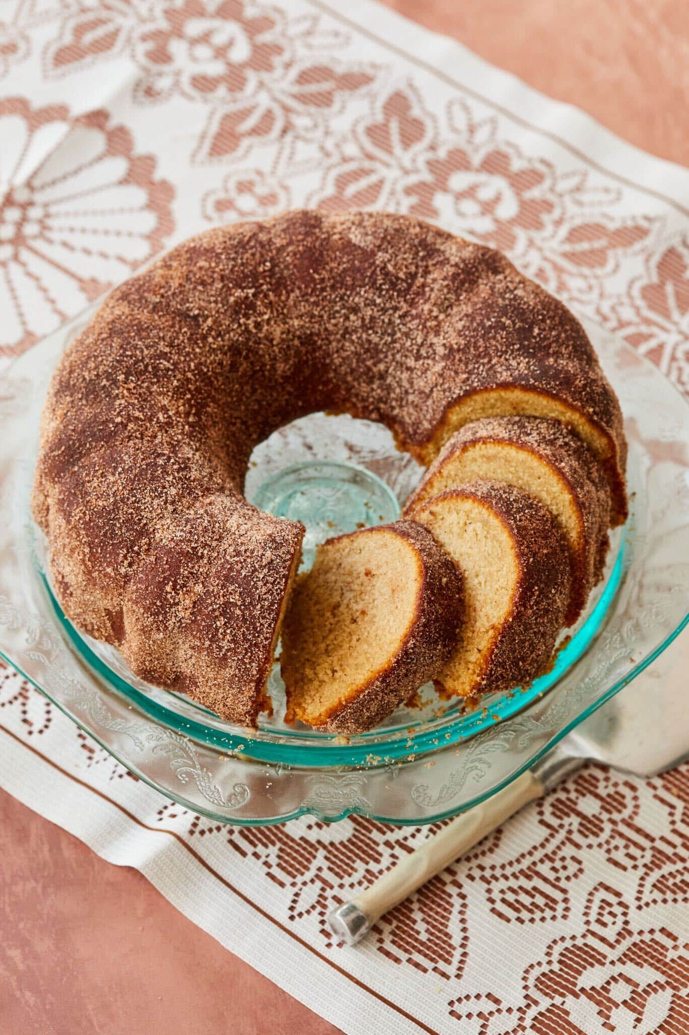 Apple Cider Donut Cake is served on a glass cake stand with a few slices cur ready for serving. The cake is shaped like a donut from the bundt cake pan and is dark golden brown on the outside with cinnamon sugar and is lightly yellow and moist on the inside. 