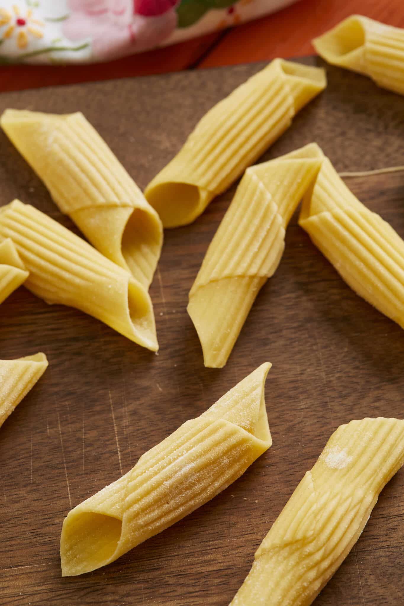 Penne pasta is drying on a wooden board. It's in a tubular-shape cut diagonally on the ends and resembles an old-fashioned writing quill that was dipped in ink. 