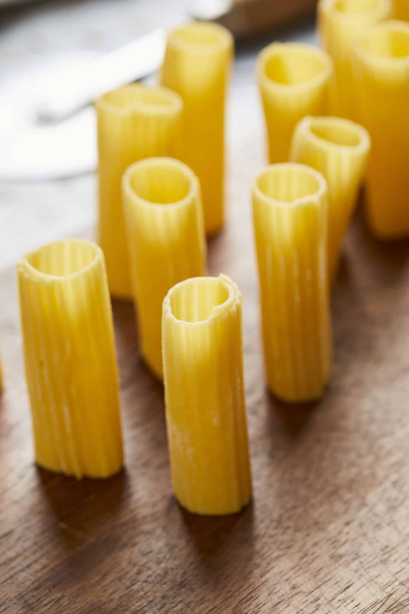Rigatoni pasta is drying upright on a wooden board. It's in yellow color, a ridged, tubular shape and about 2 inches long with squared-off ends.