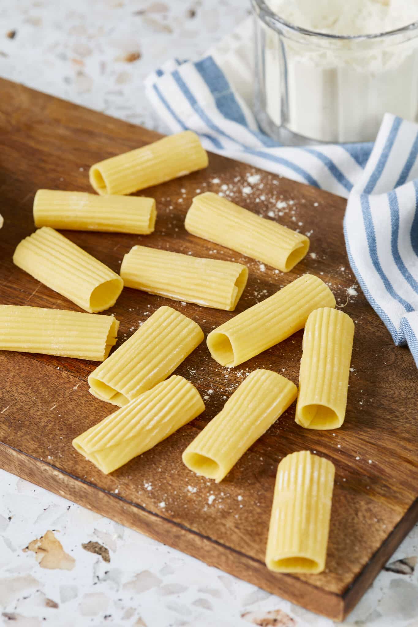 Rigatoni pasta is drying on a wooden board. It's in ridged, tubular shape and about 2 inches long with squared-off ends. It got the yellow color from semolina flour.