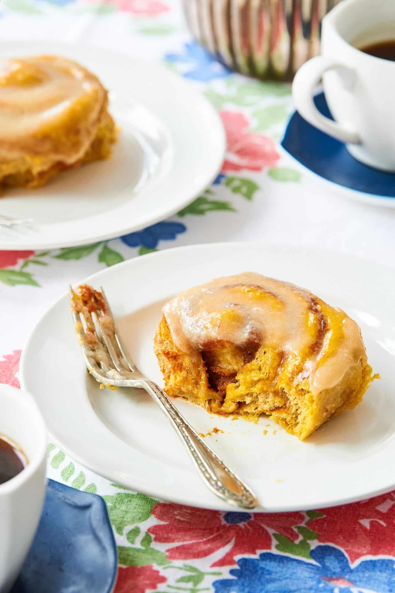 Two Pumpkin Cinnamon Rolls are served on two plates and one roll has been taken a bite. The rolls have pumpkin-infused golden orange color, are slightly crispy on the edges but very moist and soft on the inside. It's topped with cream cheese glaze and served with coffee.