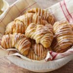 Apple Pie Crescent Rolls are baked golden brown, looking buttery, pillowy, and tender. They're drizzled with vanilla glaze and placed in a linen towel lined proofing basket.