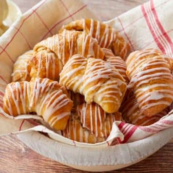 Apple Pie Crescent Rolls are baked golden brown, looking buttery, pillowy, and tender. They're drizzled with vanilla glaze and placed in a linen towel lined proofing basket.